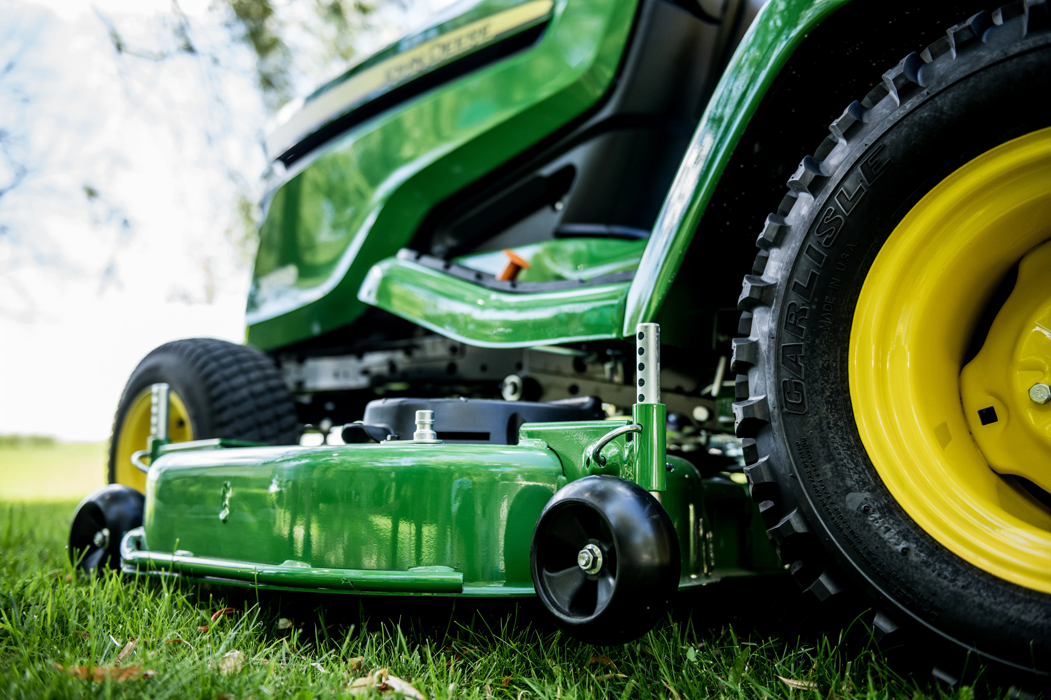 Close-up of a John Deere riding mower on grass, focusing the green mower deck, black gauge wheel, and part of the rear tire