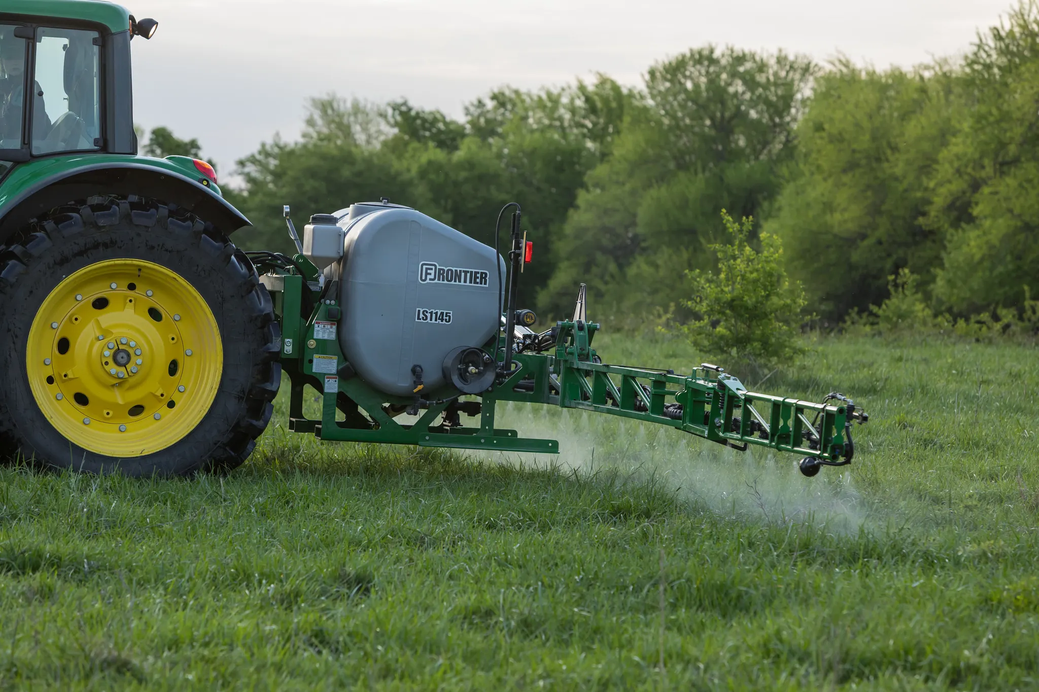 A tractor with yellow wheels pulls a mounted crop sprayer, spraying liquid over a grassy field, with trees in the background.
