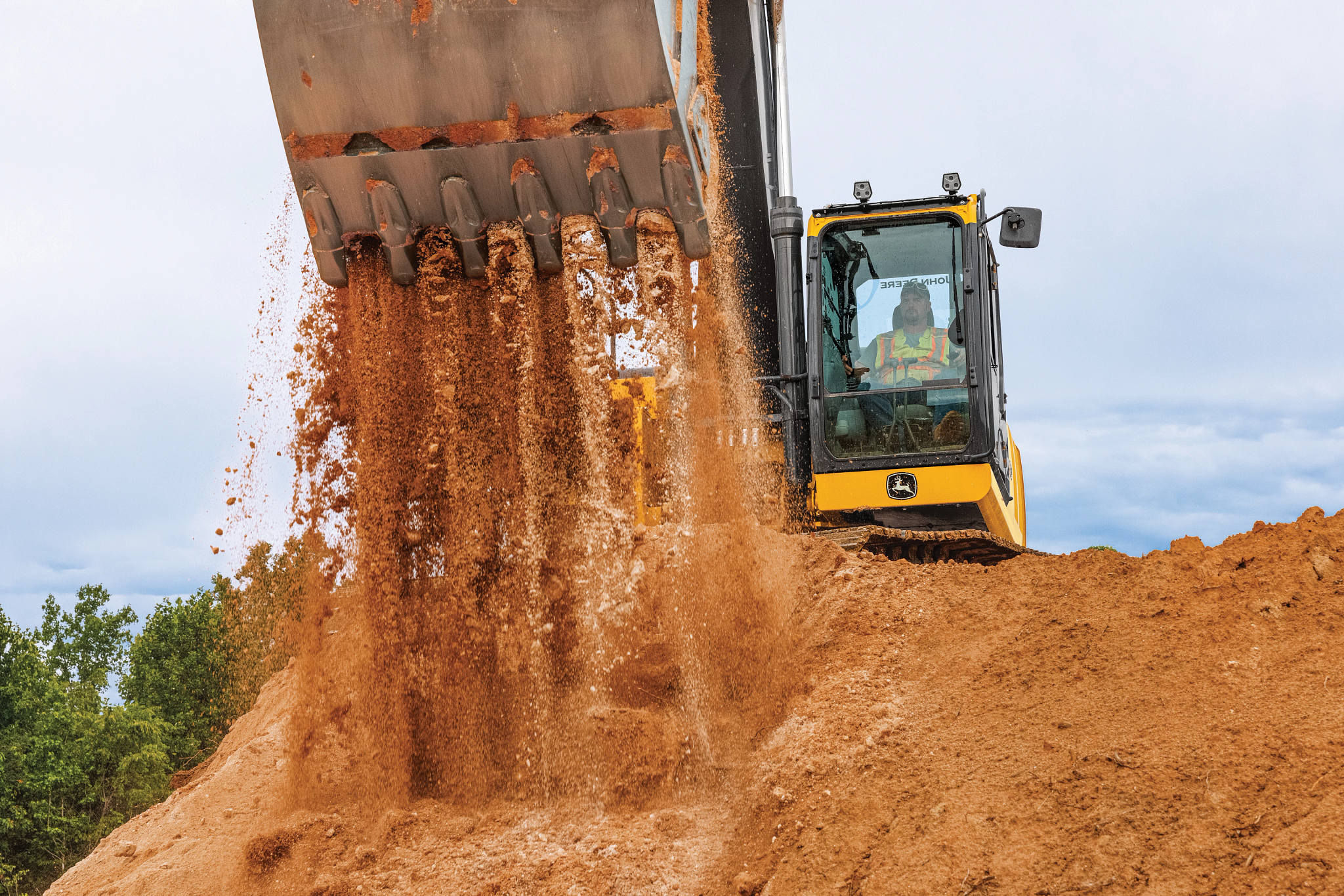 Excavator positioned atop a large dirt mound dumping soil from a raised bucket