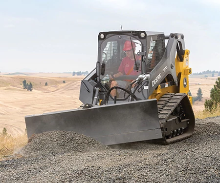 Tracked skid steer loader with a wide front blade pushing a pile of gravel on a sloped landscape, operated from an enclosed cab.