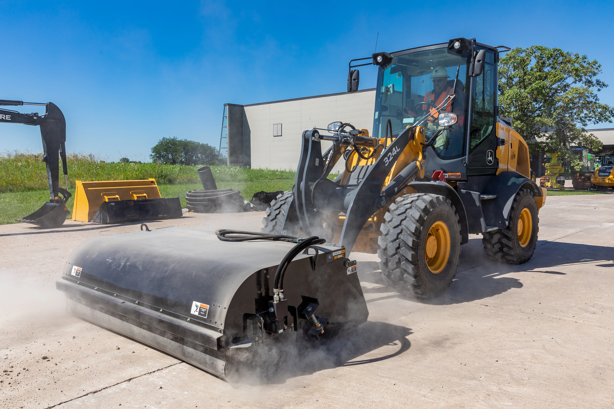 Yellow 324L compact wheel loader operating a front-mounted rotary broom, sweeping dust and debris.