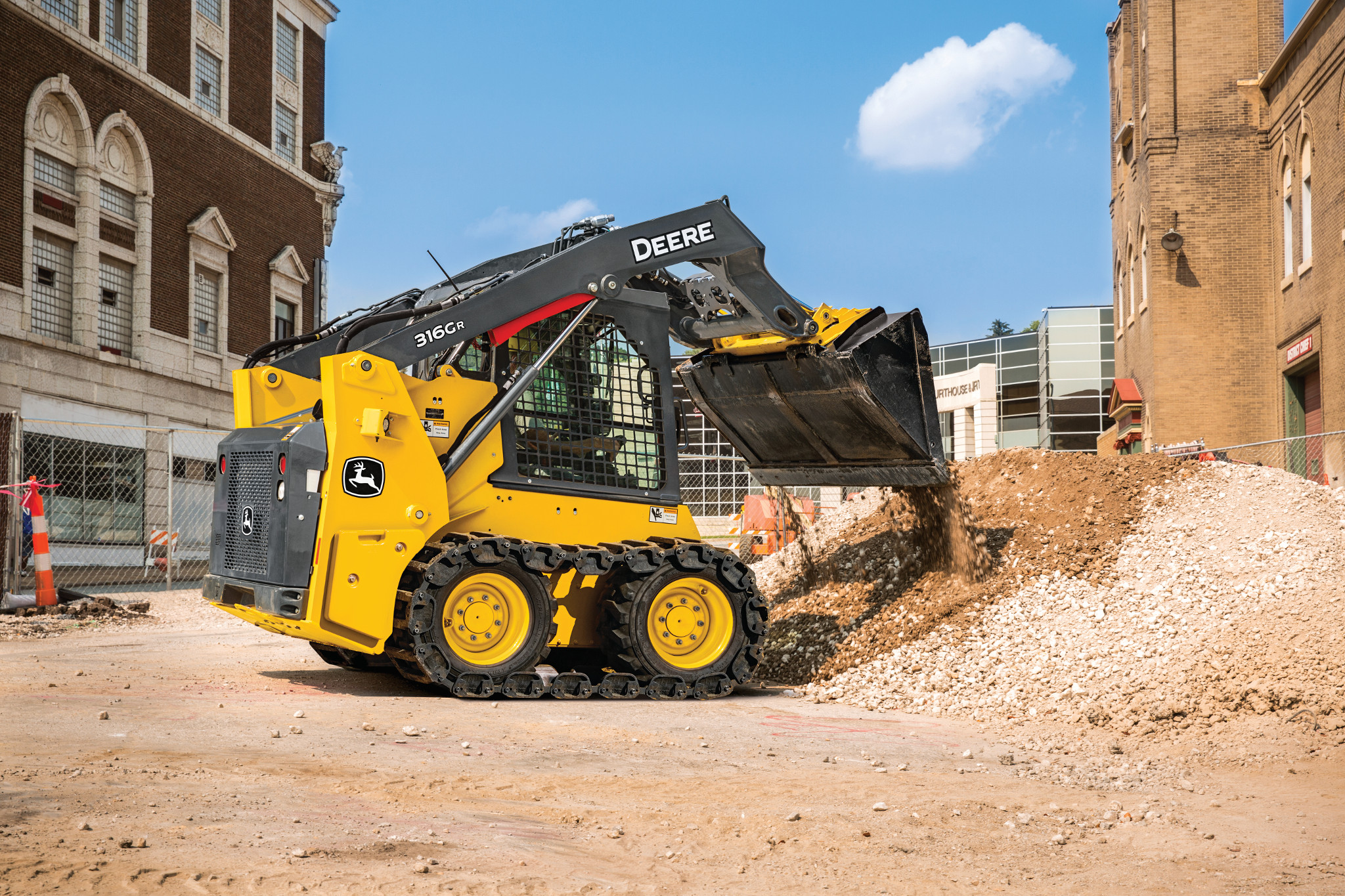 Compact track loader with a front bucket dumping gravel at a construction site between brick buildings, shown on a dirt surface under a blue sky