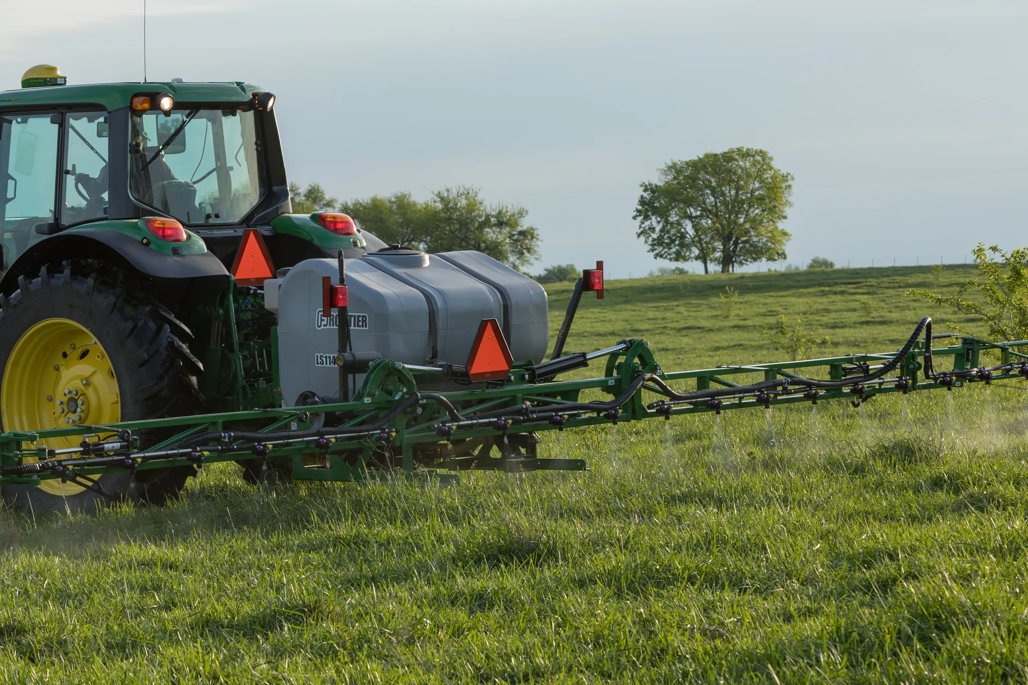 A tractor pulls a wide sprayer attachment across a grassy field, with a liquid tank mounted on the back and trees in the background under a clear sky