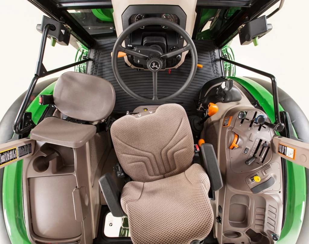 Top‑down view inside John Deere tractor cab showing fabric operator seat, steering wheel, right‑hand control console with levers and switches, cup holder, and ribbed floor mat, framed by green cab structure.