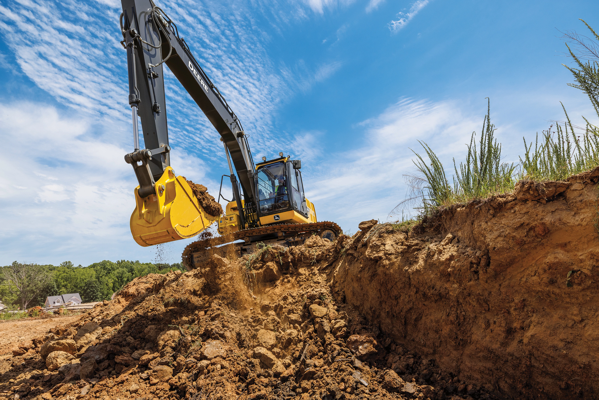 A large yellow excavator digs into a dirt embankment at a construction site, with its bucket scooping and breaking apart soil along a trench