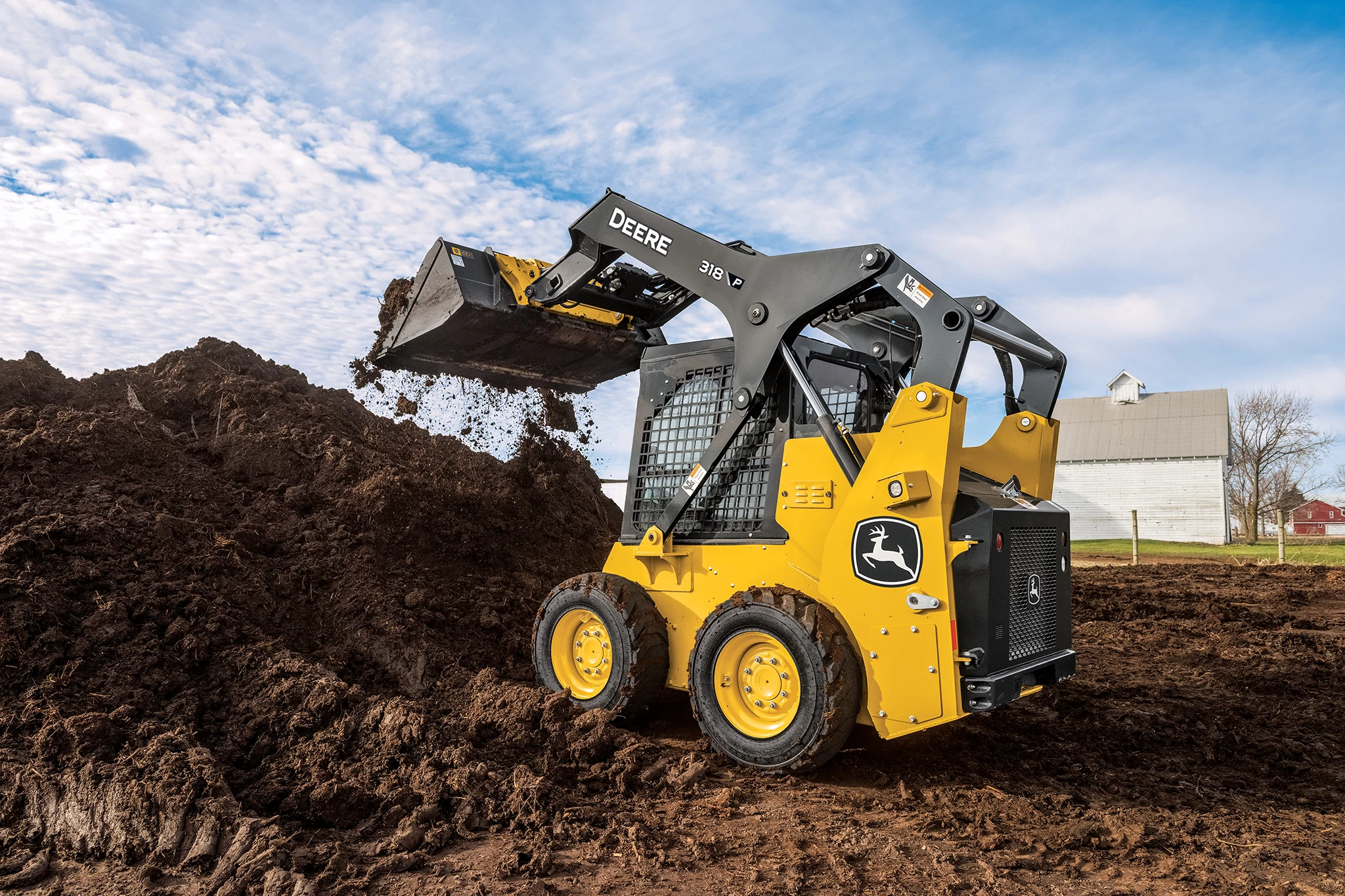 A yellow John Deere loader lifts and tips a front bucket full of dark, moist soil onto a large dirt pile at a muddy construction site.