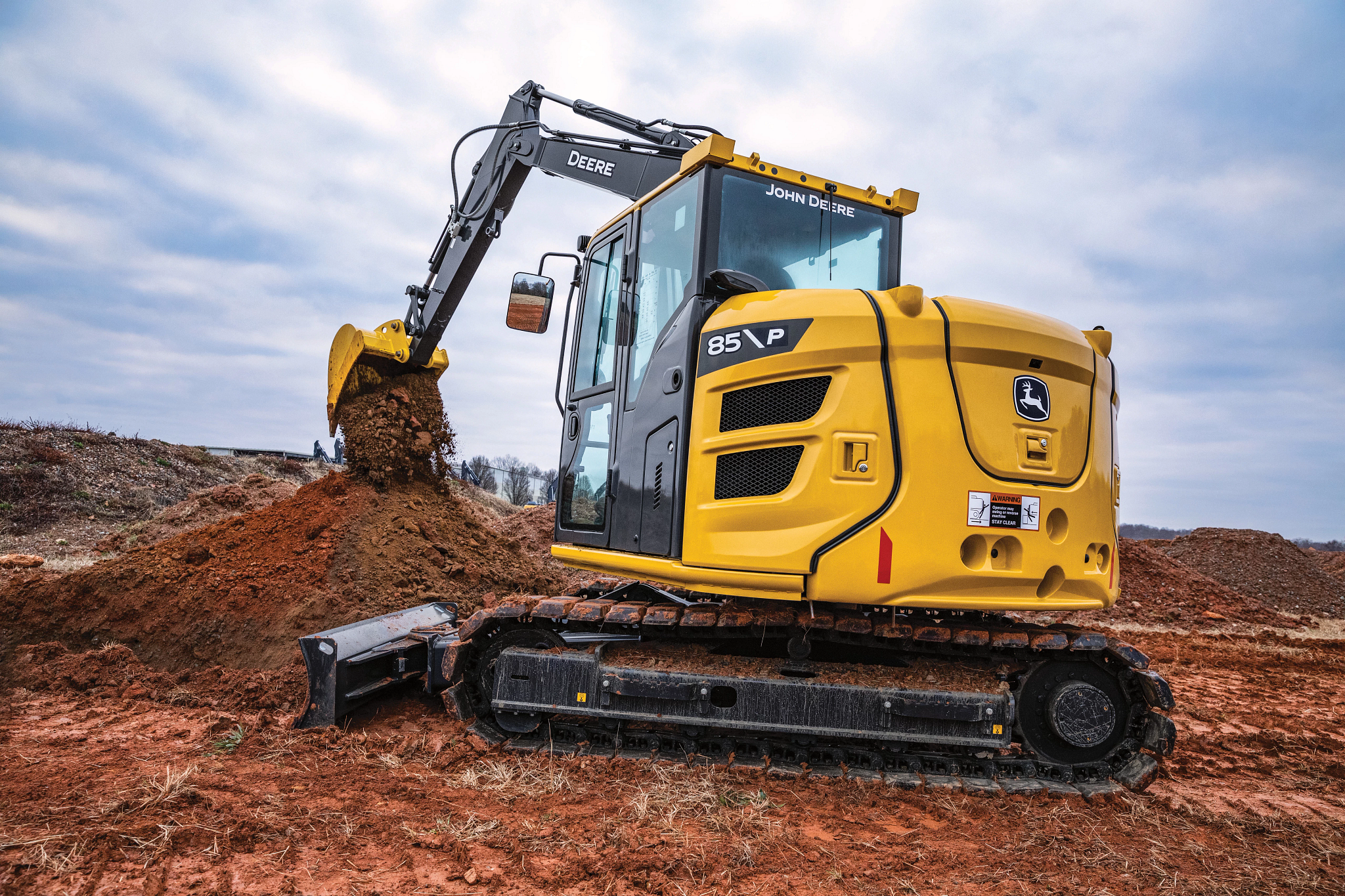John Deere 85 P compact excavator working on a dirt construction site, featuring a yellow and gray body, enclosed cab, and metal tracks
