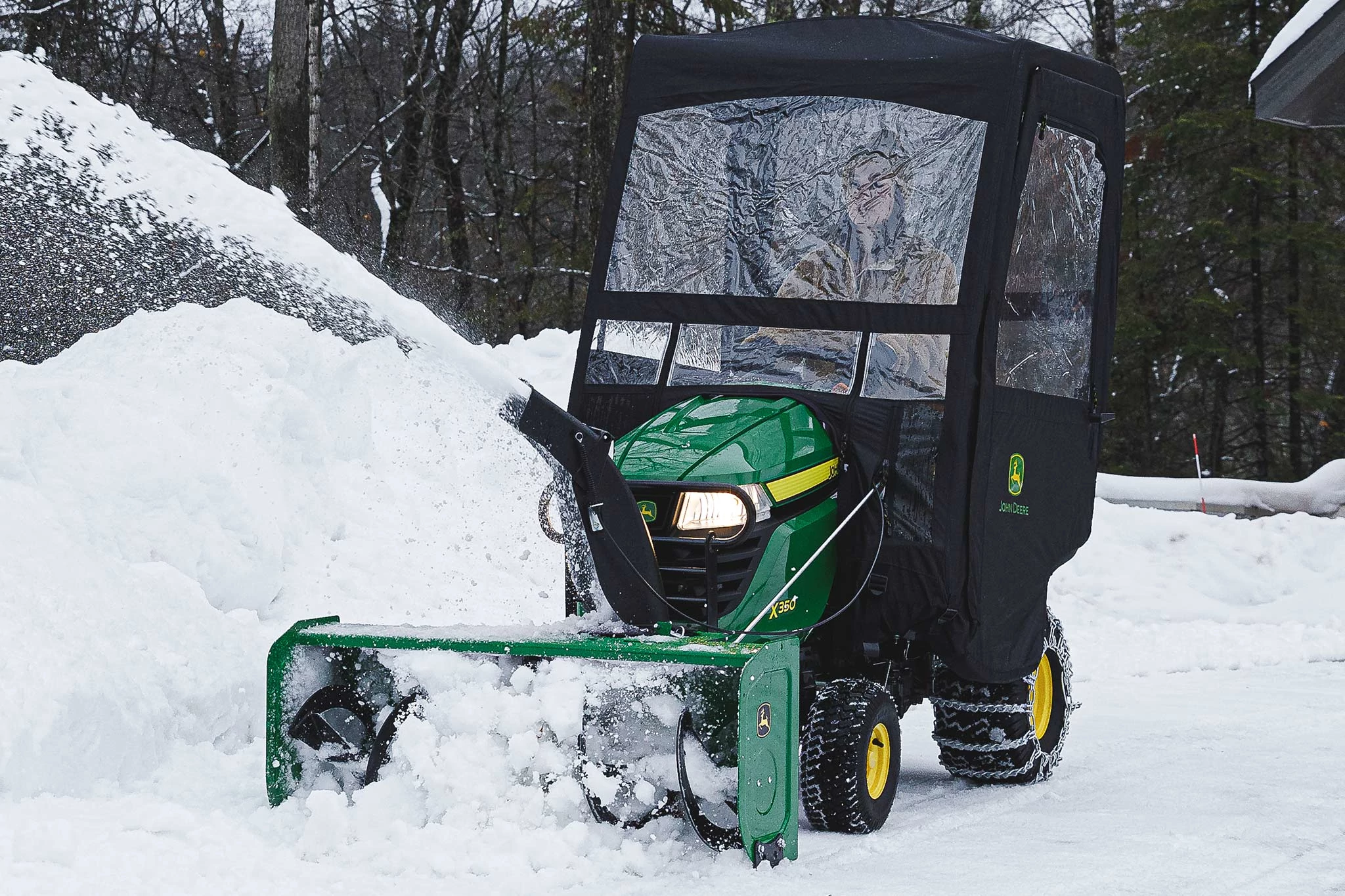 Front left view of an operator inside a weather enclosure on an X350 Tractor clearing a snowy driveway with a 44-in. Snow Blower and tire chains