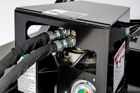 Close-up of a black metal hydraulic power unit housing with two connected hydraulic hoses, fittings, and warning labels, shown against a neutral background