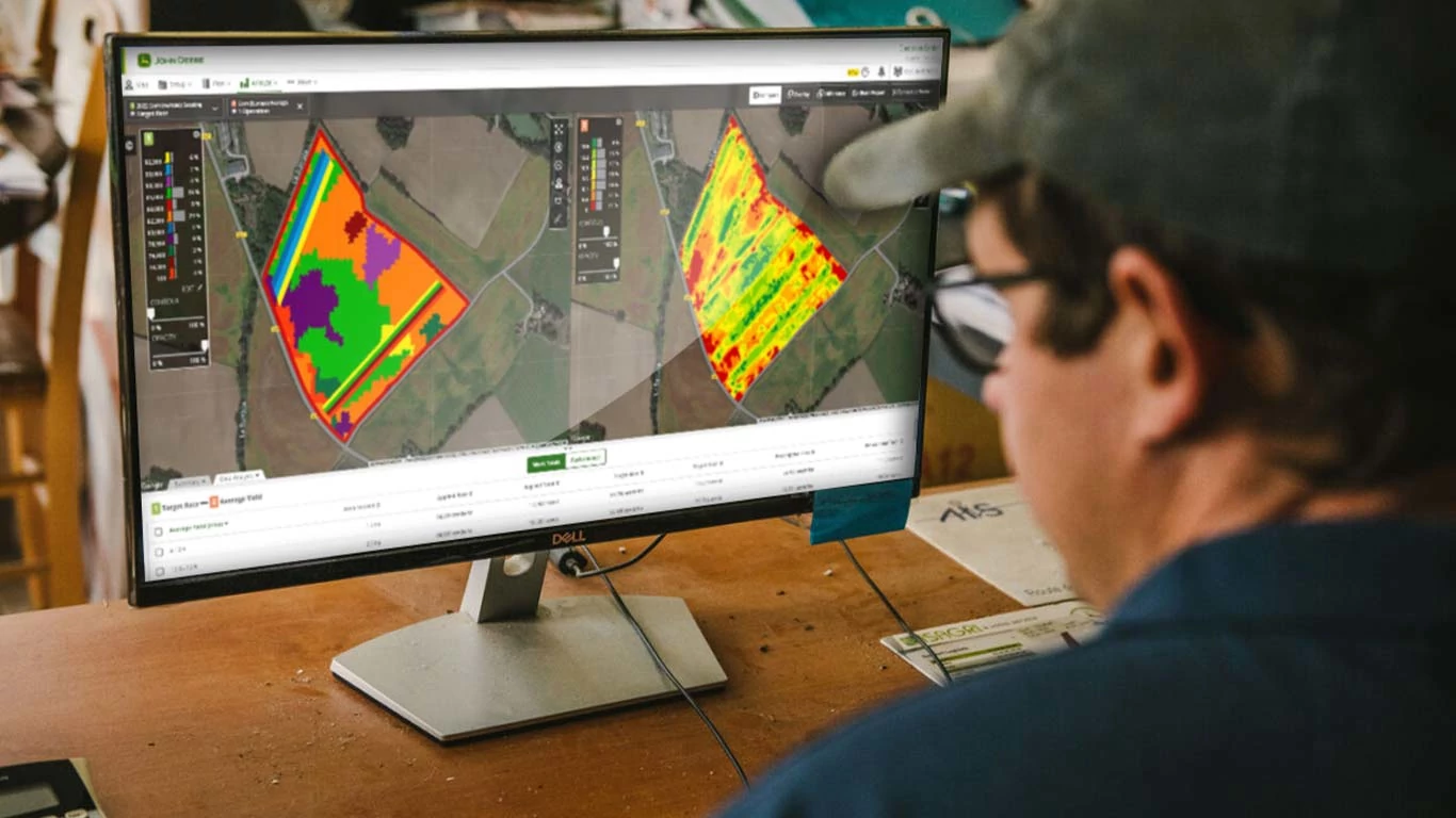 Person viewing John Deere Operations Center maps on a desktop monitor, showing field analysis layers and crop performance data