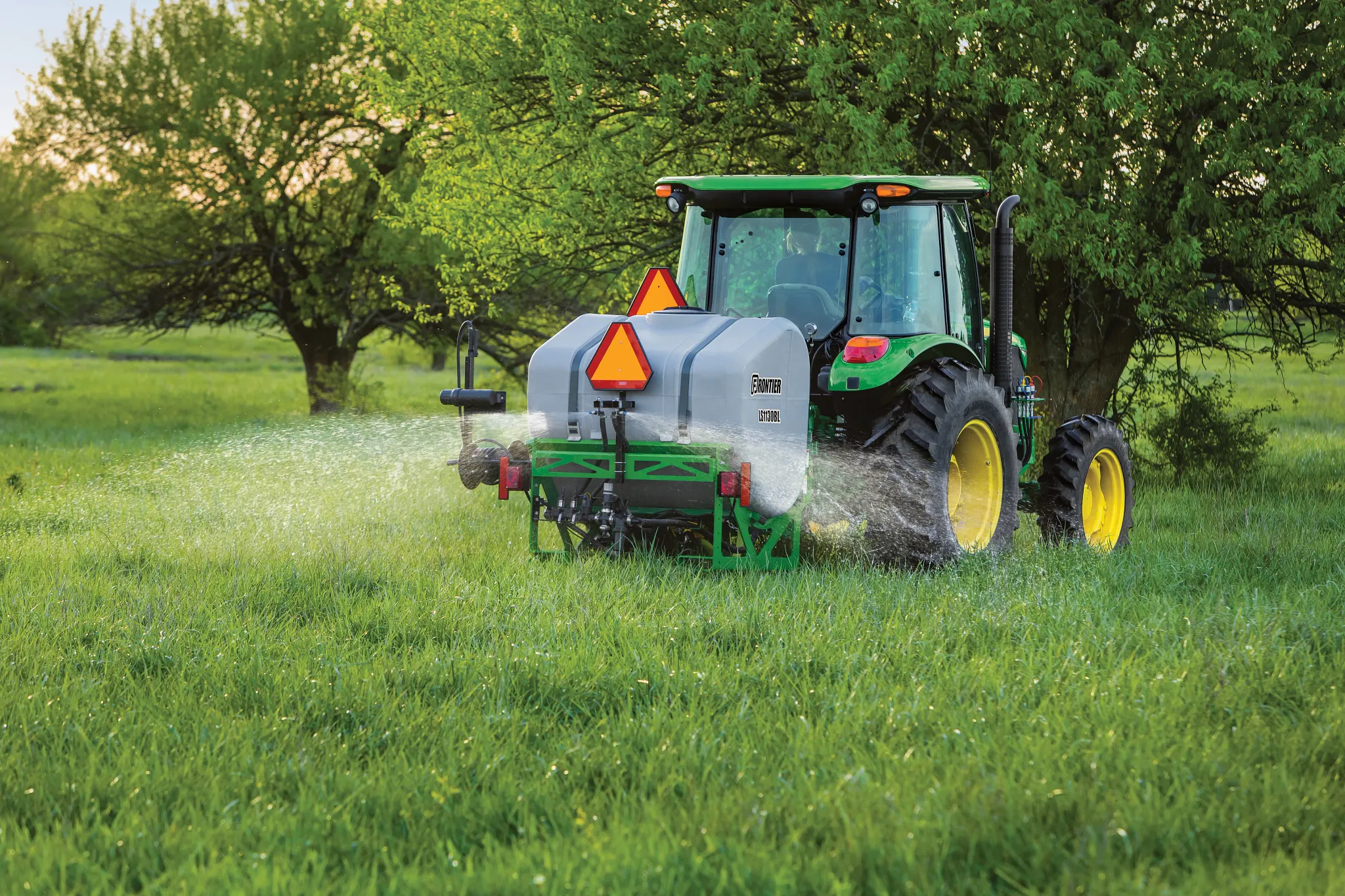 A tractor drives through a grassy field, spraying liquid from a tank-mounted applicator on the back, with trees in the background on a sunny day.