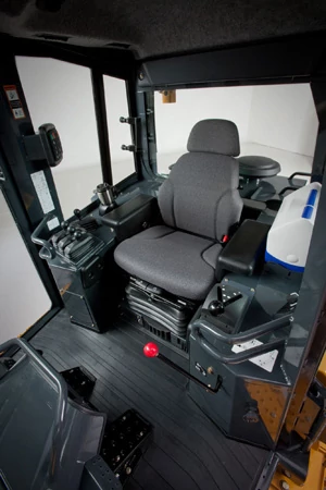 Interior of a crawler loader operator cab showing a padded seat with armrests, control panels, joysticks, levers, foot pedals, and storage areas