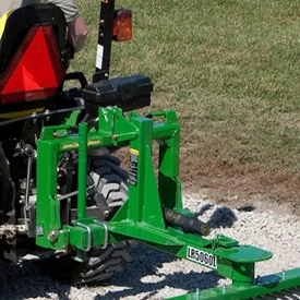 A green landscape rake with a drawbar-style hitch rests on gravel and grass, showing a metal frame, and hydraulic components, while attached to the rear of a tractor