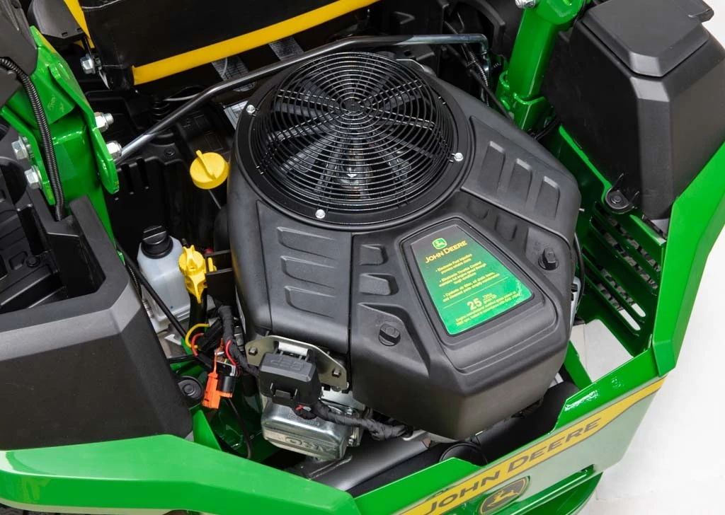 Top view of a John Deere zero‑turn mower engine compartment with black engine cover, cooling fan grille, yellow oil cap, and green mower frame, John Deere label visible on the engine cover.
