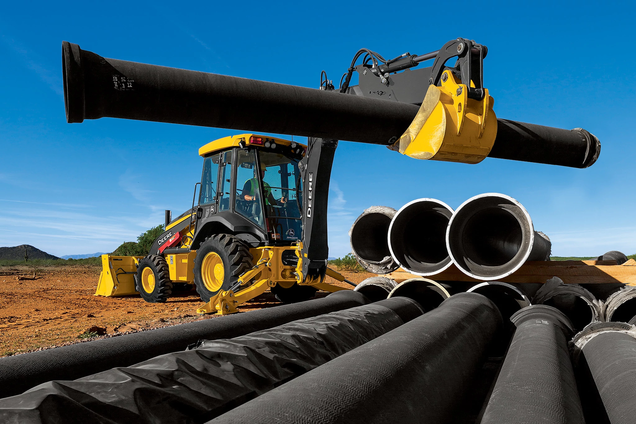 310 P-Tier Backhoe lifting a long black pipe at a dusty outdoor worksite