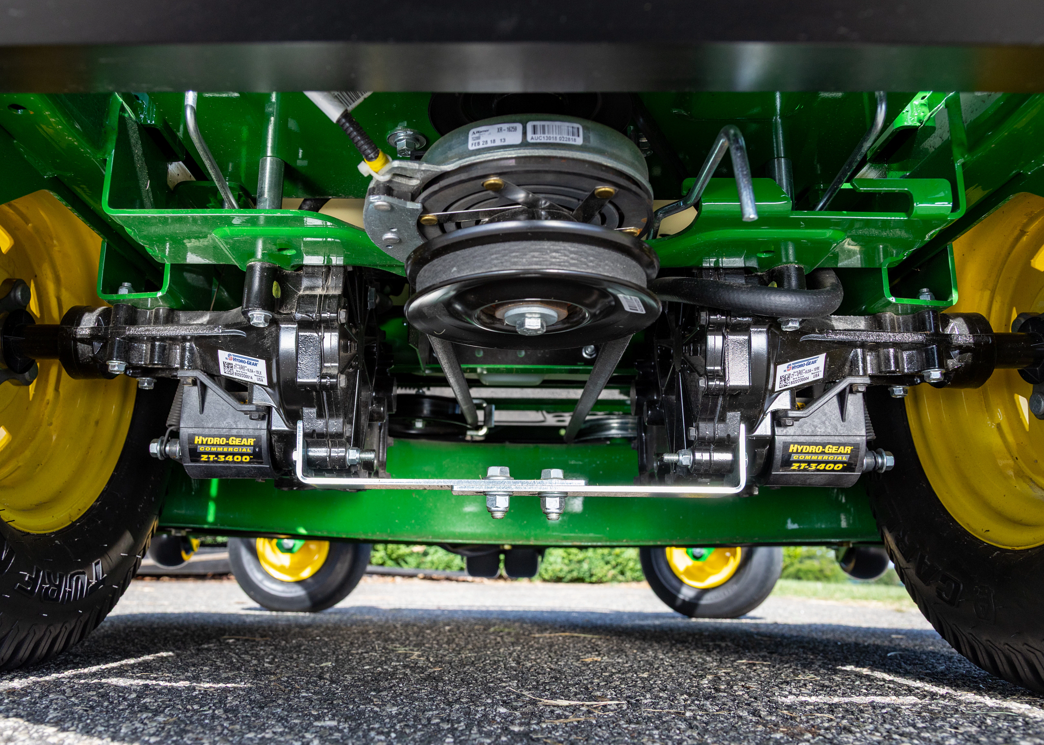 Underside view of a riding mower or tractor showing the drive system, including twin hydrostatic transmissions, belt and pulley assembly, axle components, and yellow wheels.
