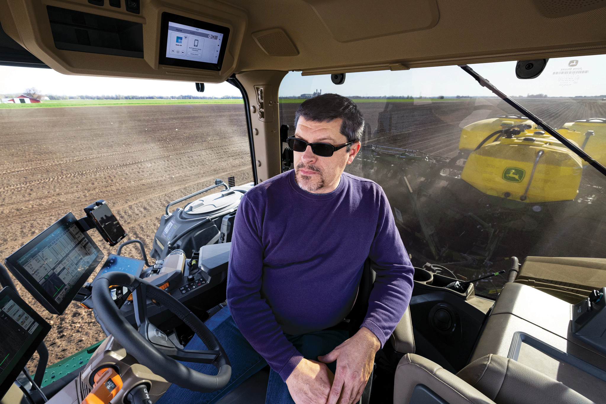 Person in sunglasses seated inside a modern tractor cab in a large cultivated field showing a steering wheel, monitors, and equipment.