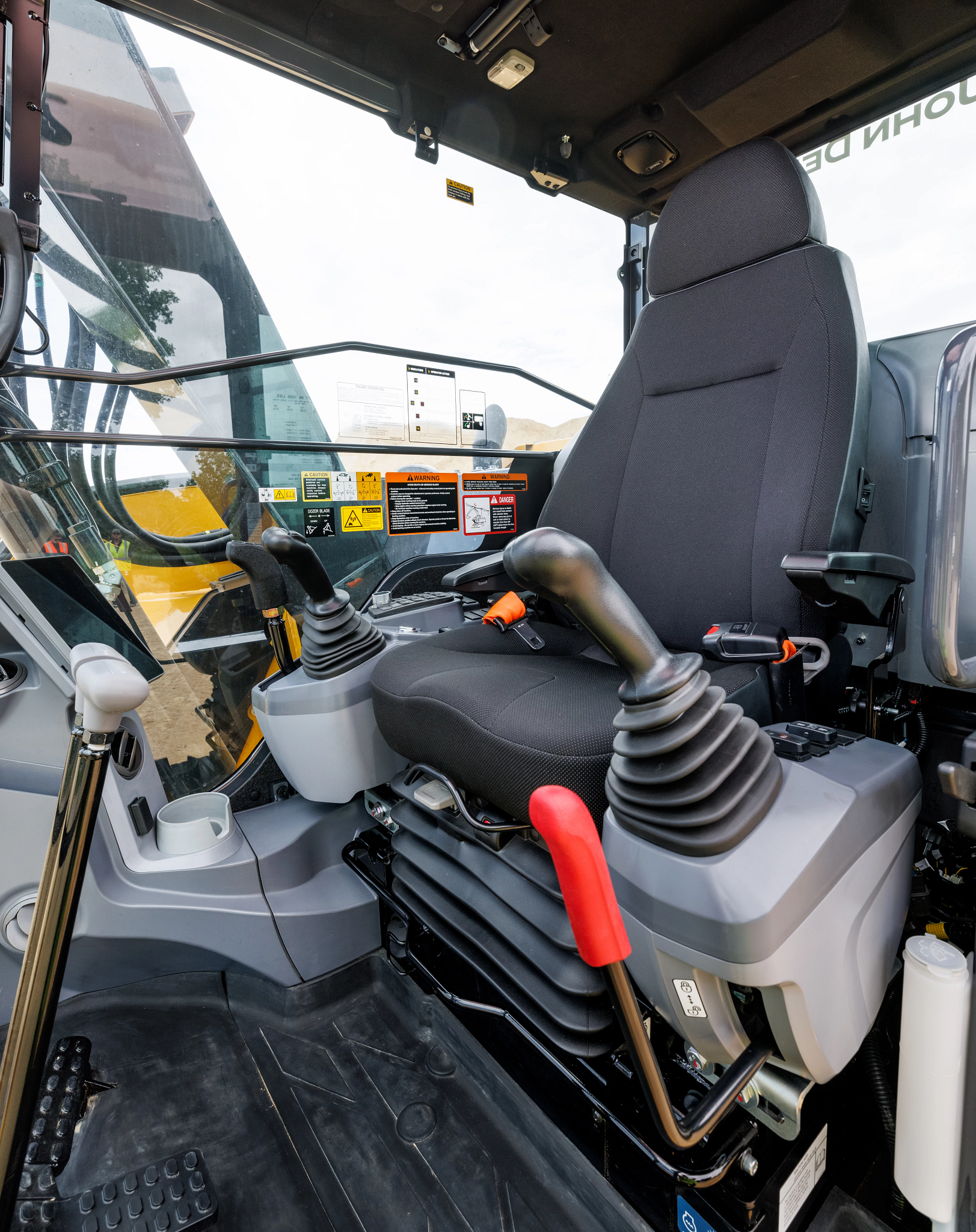 Interior of construction equipment cab showing a black operator seat with armrests, dual joystick controls, foot pedals, and safety labels.
