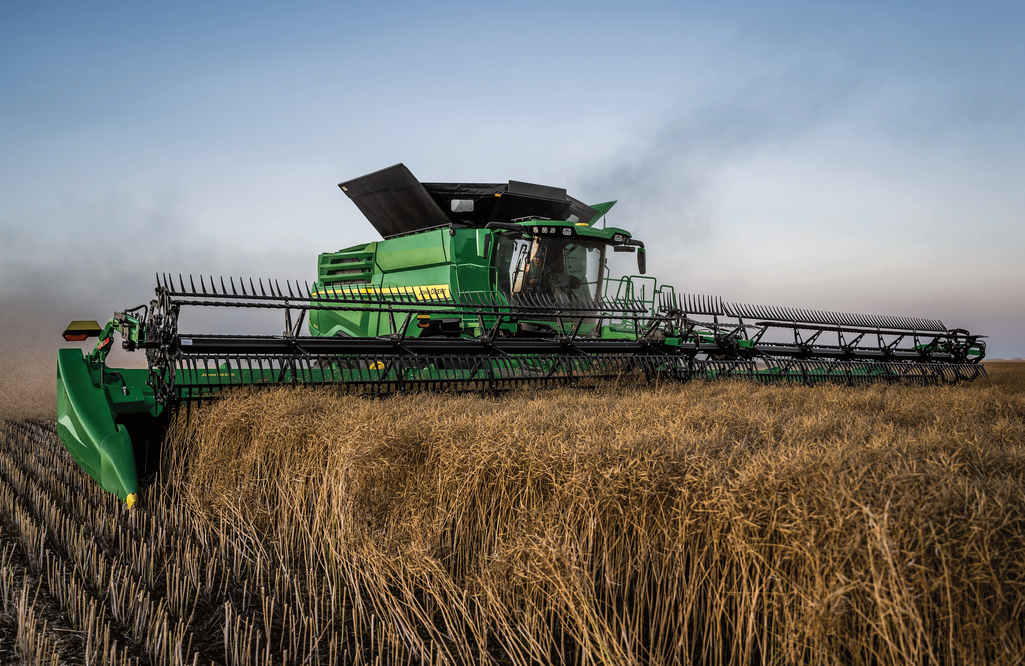 Green combine harvester cutting through a mature field of dry grain with a wide header, leaving short stubble behind, as dust rises lightly under a pale sky with a distant, flat horizon