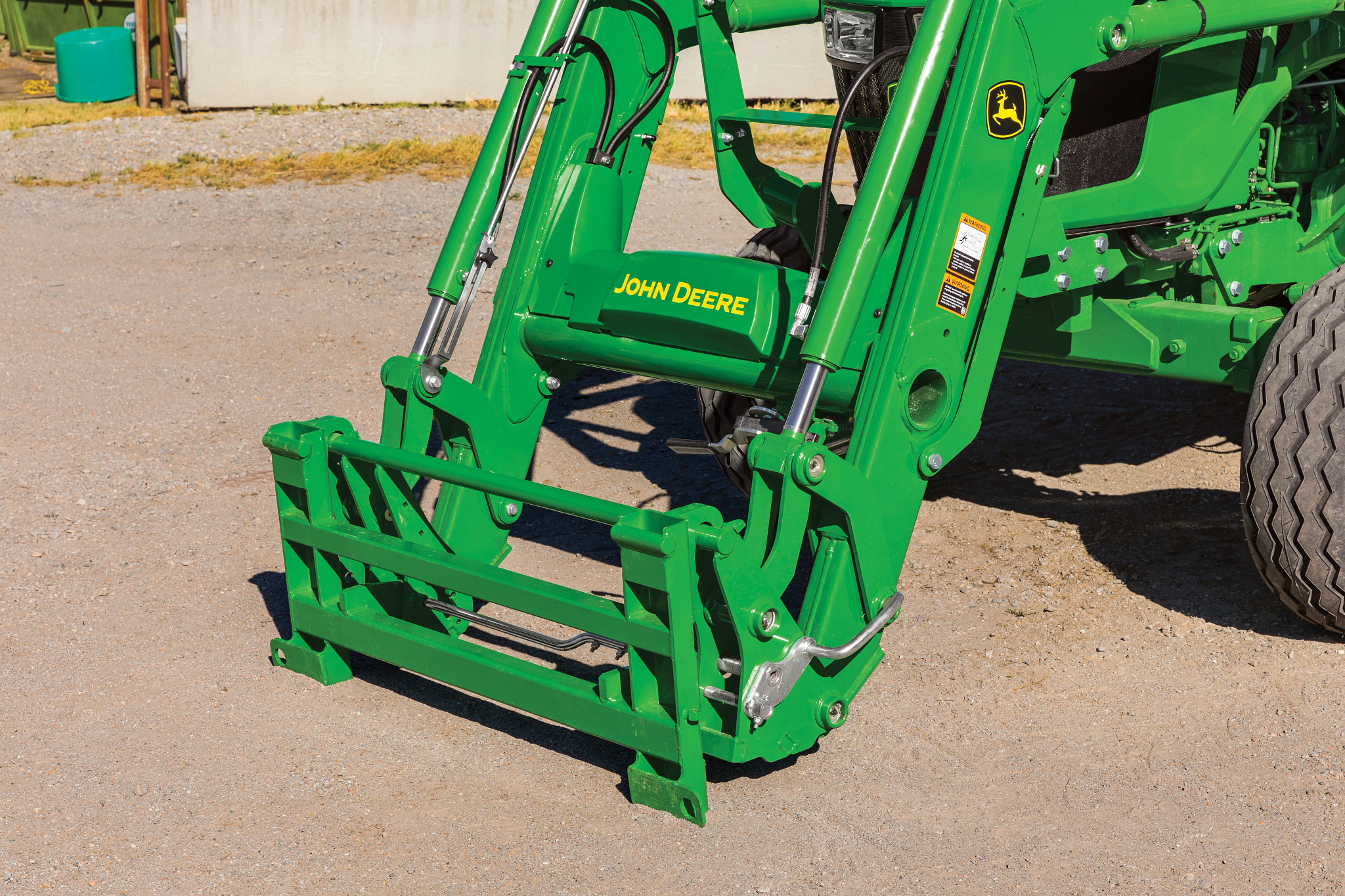 A close-up of a green front loader attachment showing the hydraulic arms, hoses, and quick‑attach carrier adapter positioned over a gravel surface