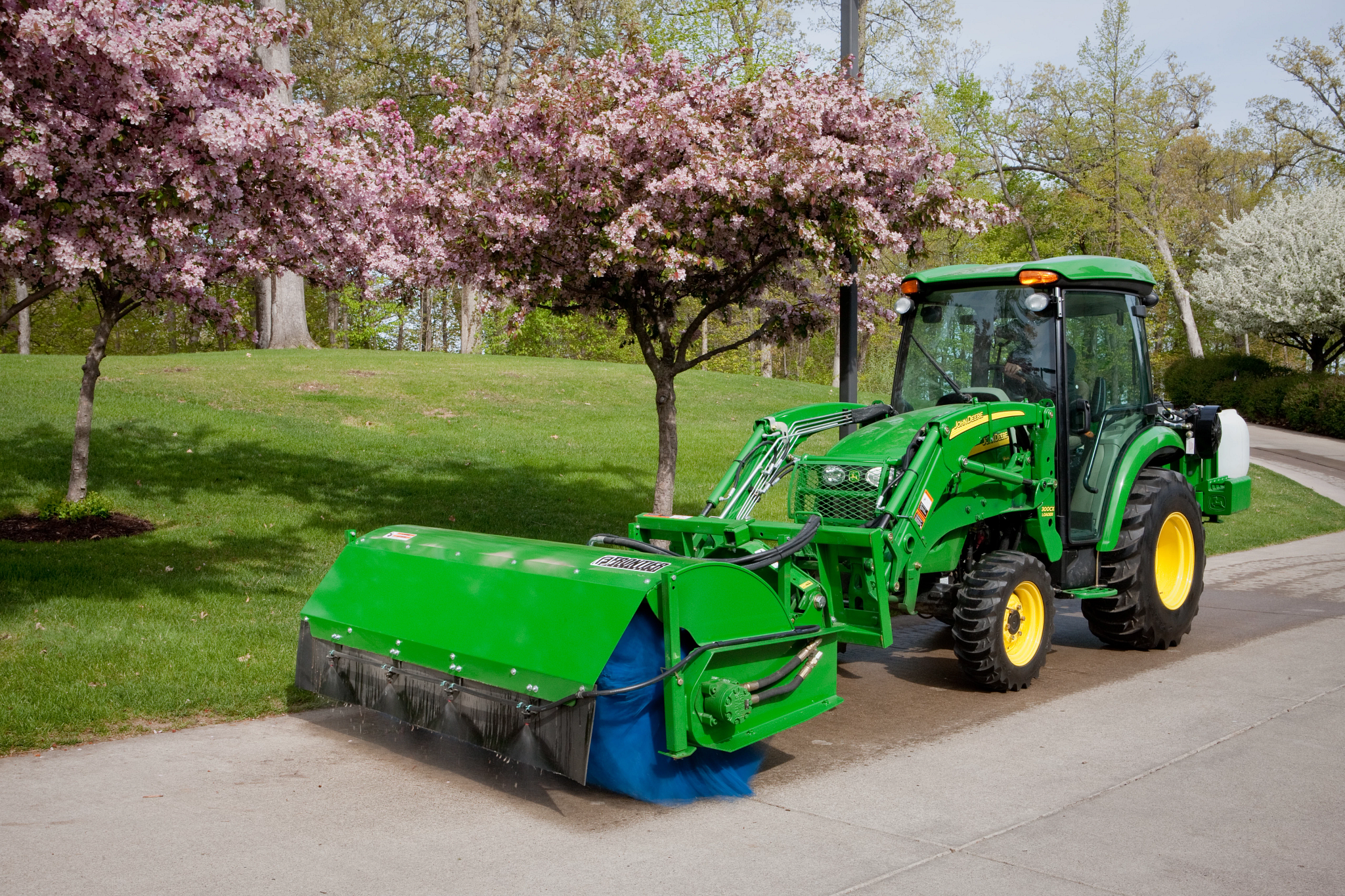 Front left view of a SW21 front mount rotary broom on a 300CX carrier loader using the optional dust suppression system along a road lined with flowering trees