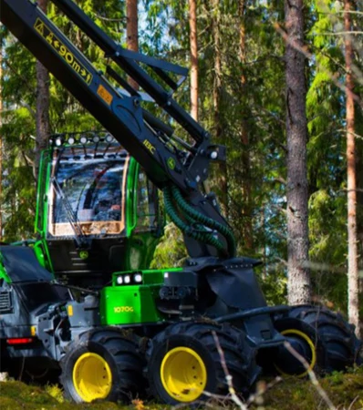 A green forestry machine with multiple large yellow wheels and a long articulated boom operates among tall trees in a wooded forest setting