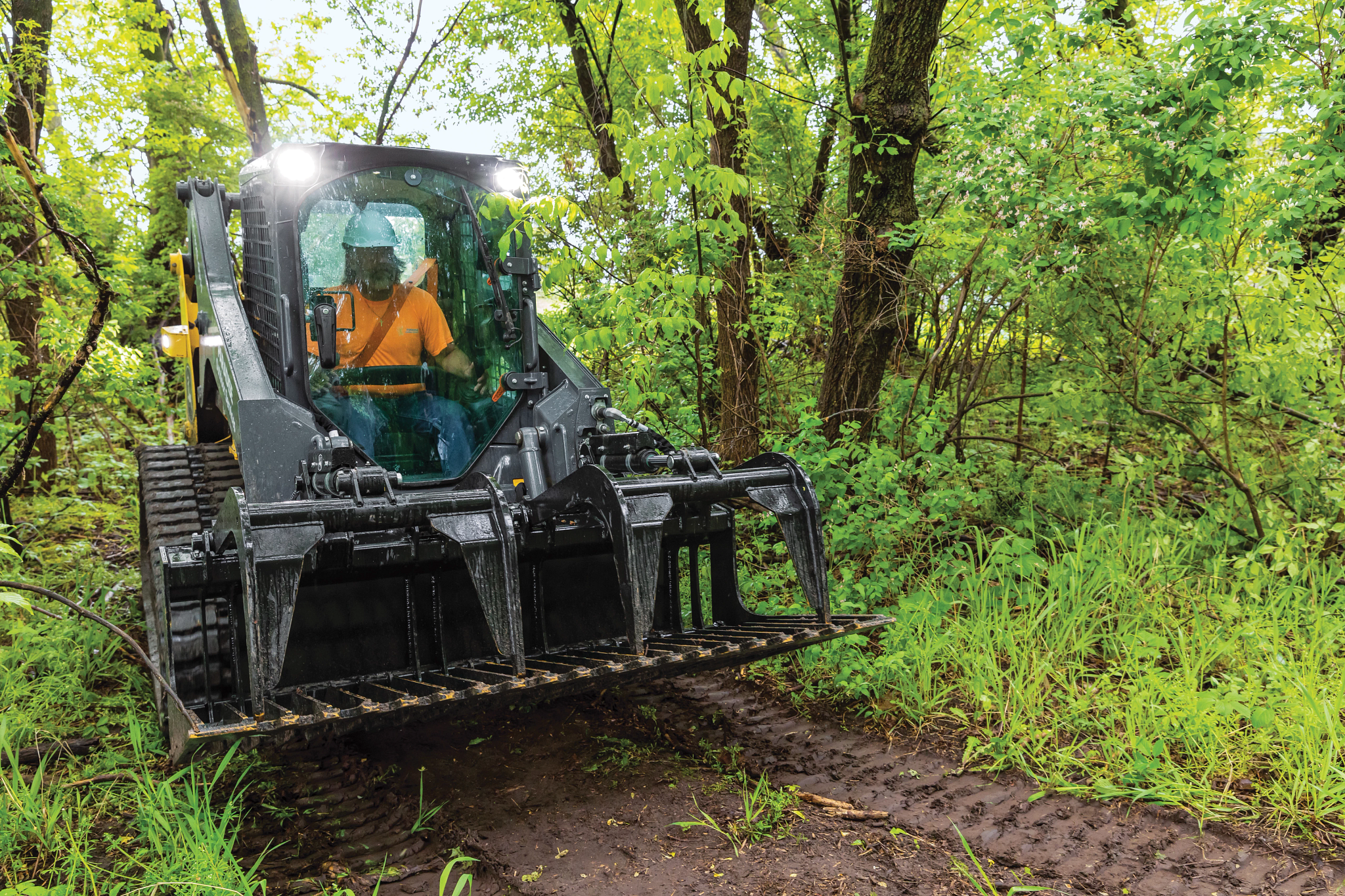 Tracked compact loader with a front grapple attachment driving along a narrow dirt path in a wooded area with dense green vegetation