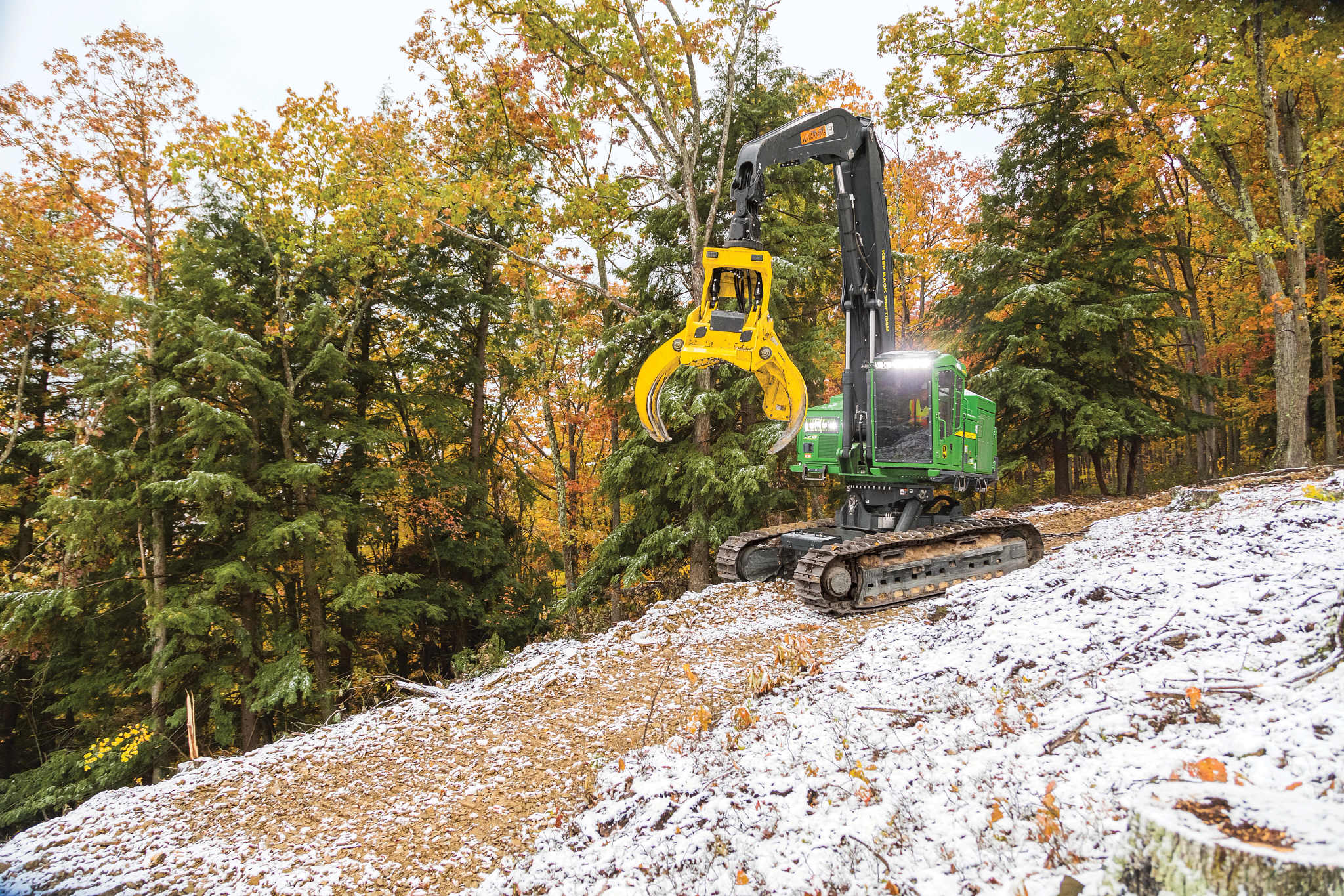John Deere forestry machine uses a grapple to handle logs on a snowy slope, surrounded by autumn-colored trees and dense forest, with tracked undercarriage navigating uneven terrain.
