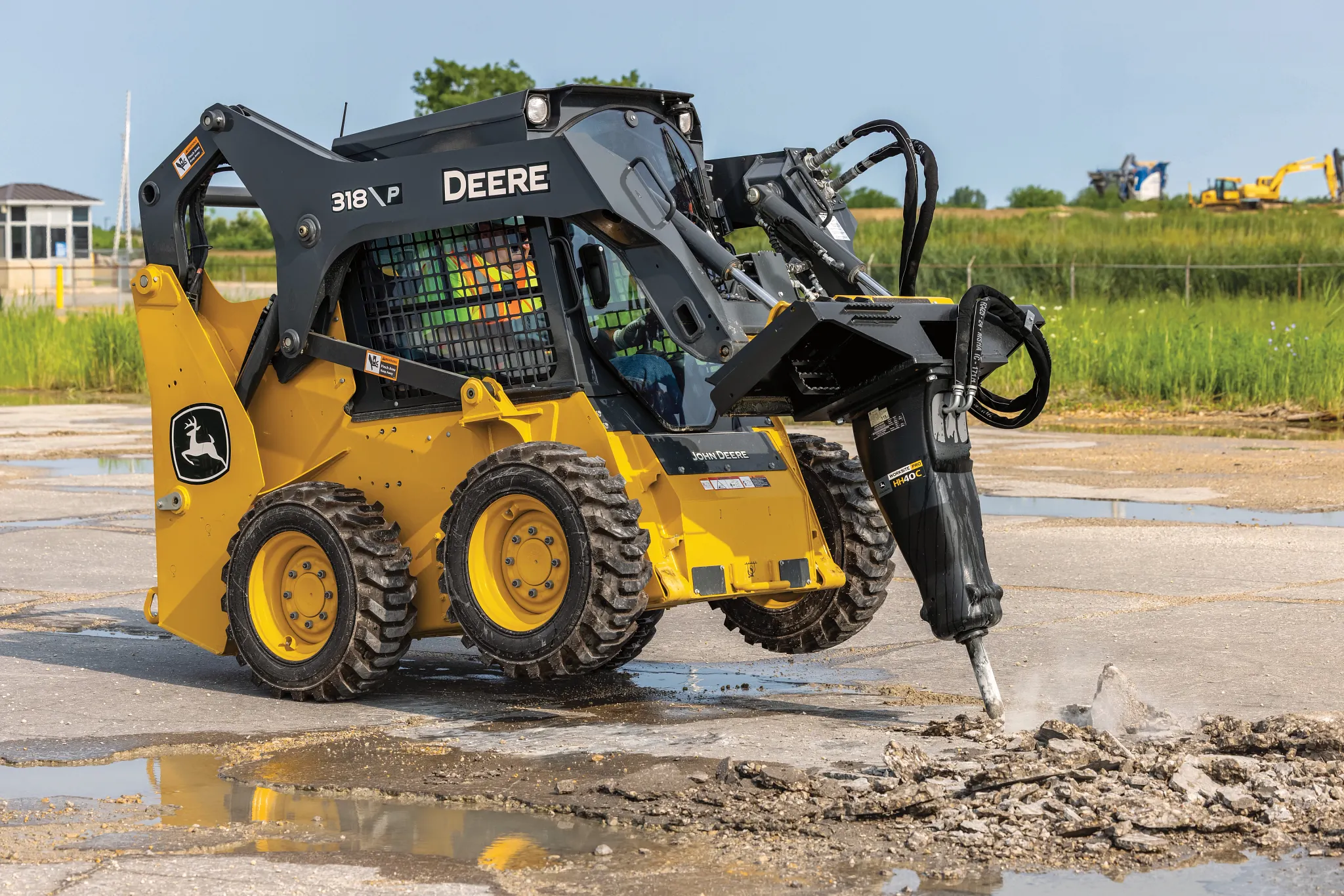 A 318 P-Tier skid steer operates on a paved surface, using a mounted hydraulic breaker attachment to break up concrete.