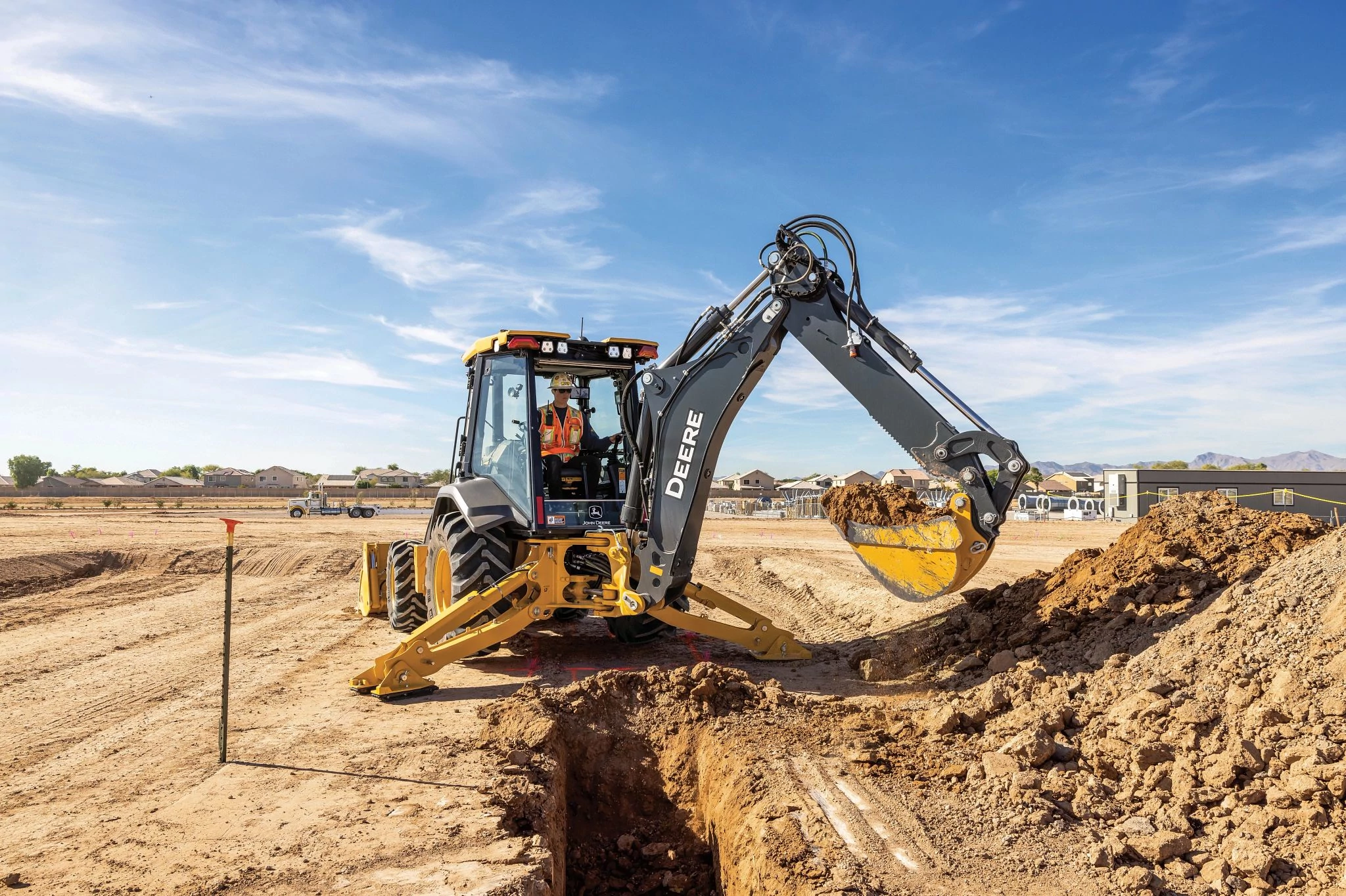 A yellow John Deere backhoe loader excavates a trench at a sandy construction site, with the machine’s arm extended and its bucket lifting a load of soil