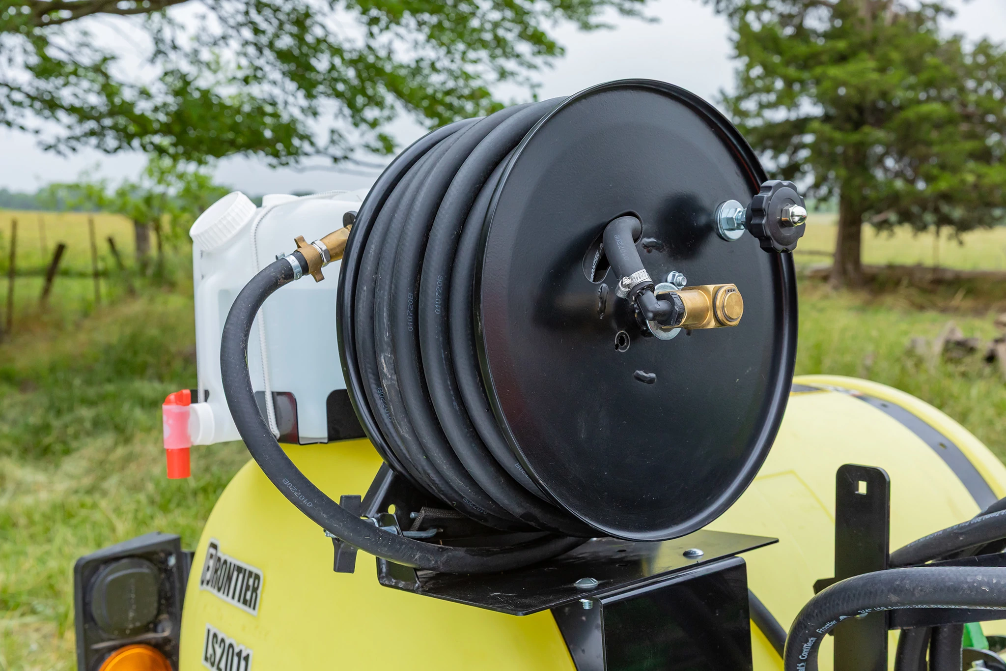 Close-up of a hose reel mounted on a yellow agricultural sprayer, featuring a coiled black hose and fittings, with a grassy field and trees in the background.