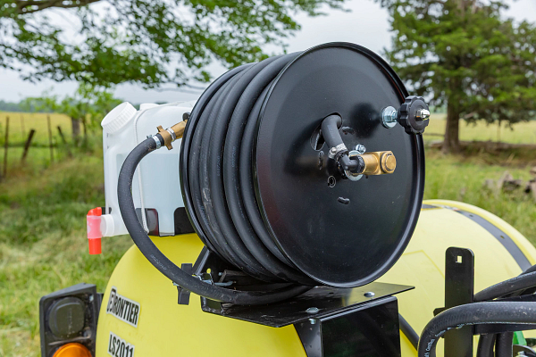 Close-up of a hose reel mounted on a yellow agricultural sprayer, featuring a coiled black hose and fittings, with a grassy field and trees in the background.