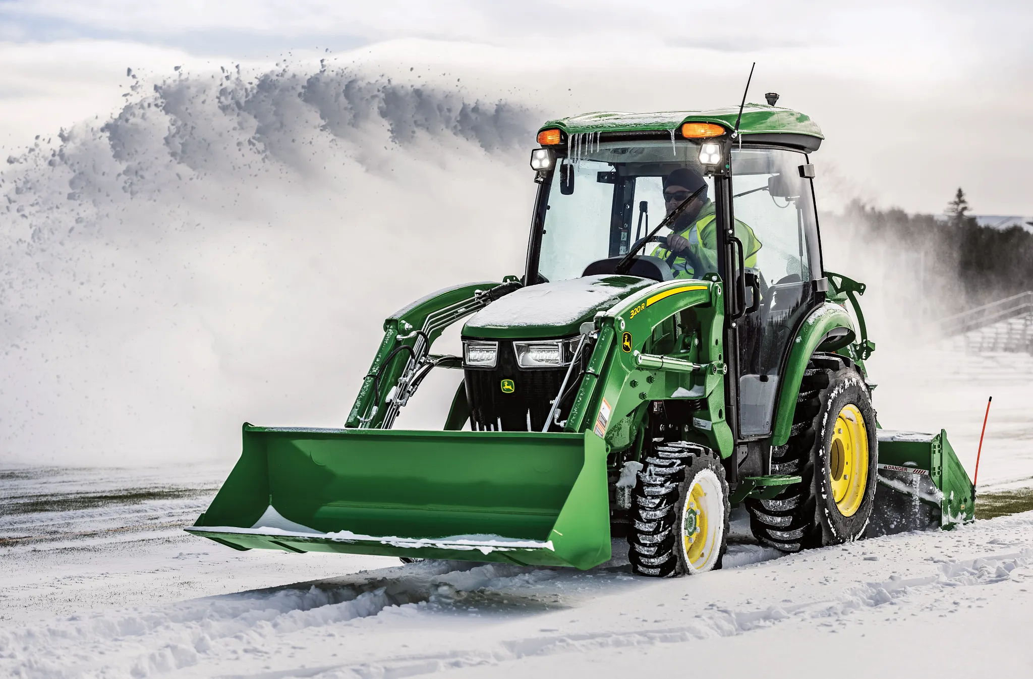 Front left view of a green 3046R compact utility tractor with 320R loader attachment snow blowing a road with rear-attached SB1164P snowblower