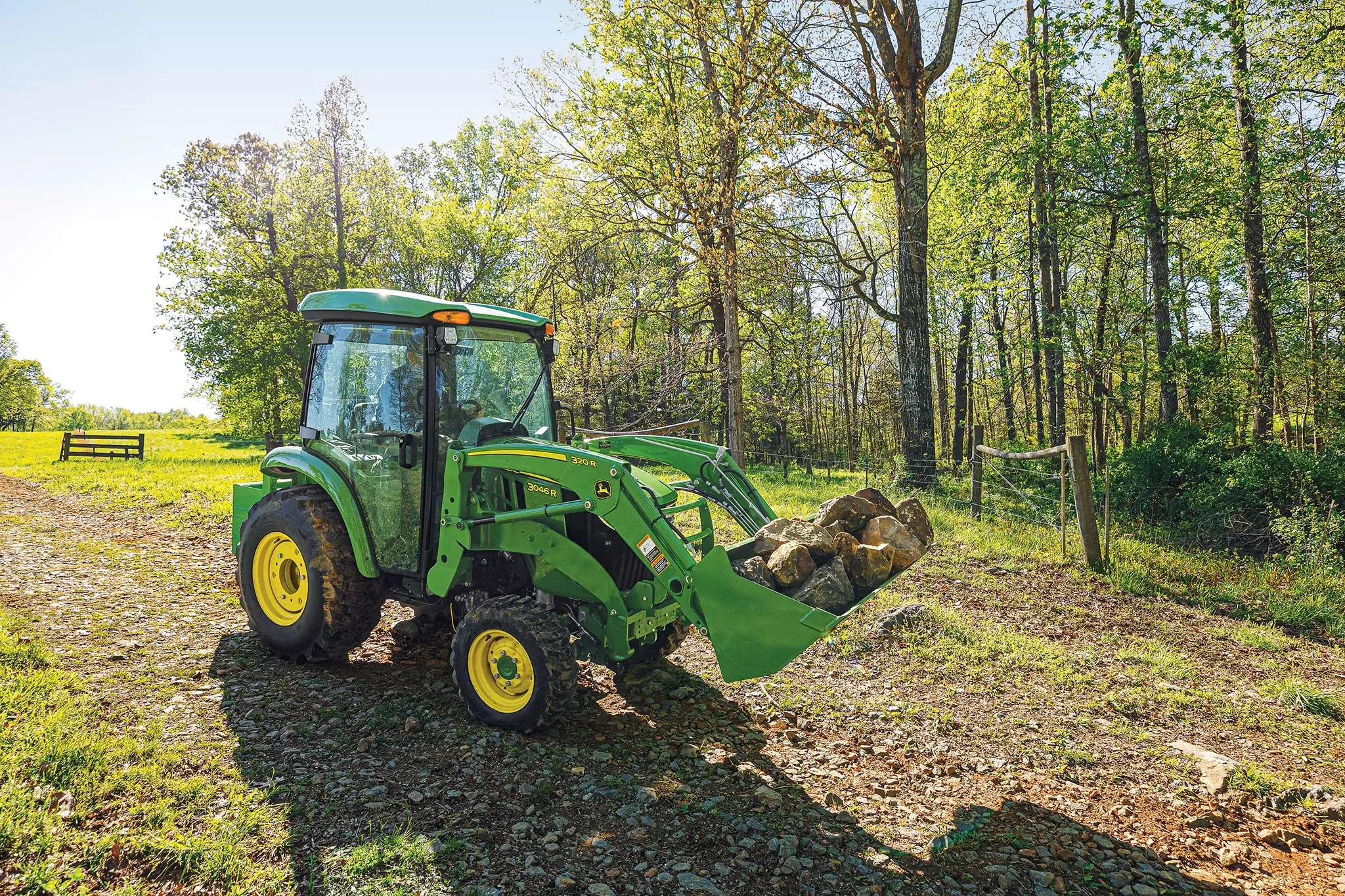 Front right view of a 3046R compact utility tractor with front-attached 320R loader hauling boulders across a dirt path near the woods