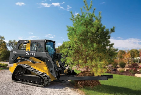 Side right view of a 329E compact track loader with attached NF44 nursery fork carrying a white pine tree