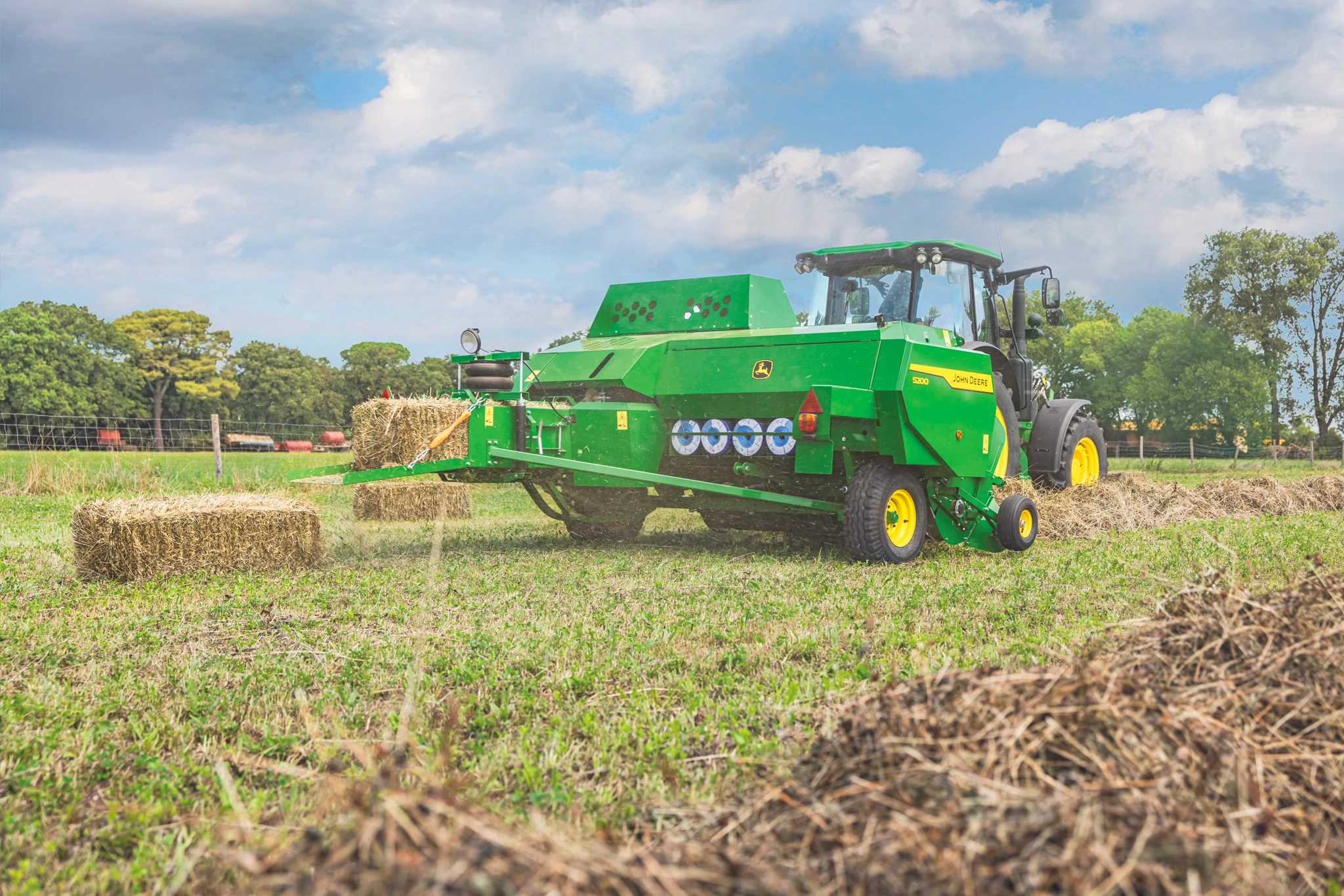 Green tractor pulling an S200 square baler across a harvested field, forming rectangular hay bales. Fresh bales lie on the ground, with trees and a fence in the background beneath a partly cloudy blue sky.