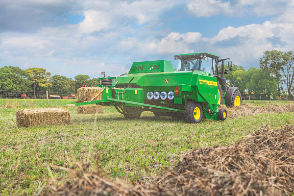 Green tractor pulling an S200 square baler across a harvested field, forming rectangular hay bales. Fresh bales lie on the ground, with trees and a fence in the background beneath a partly cloudy blue sky.
