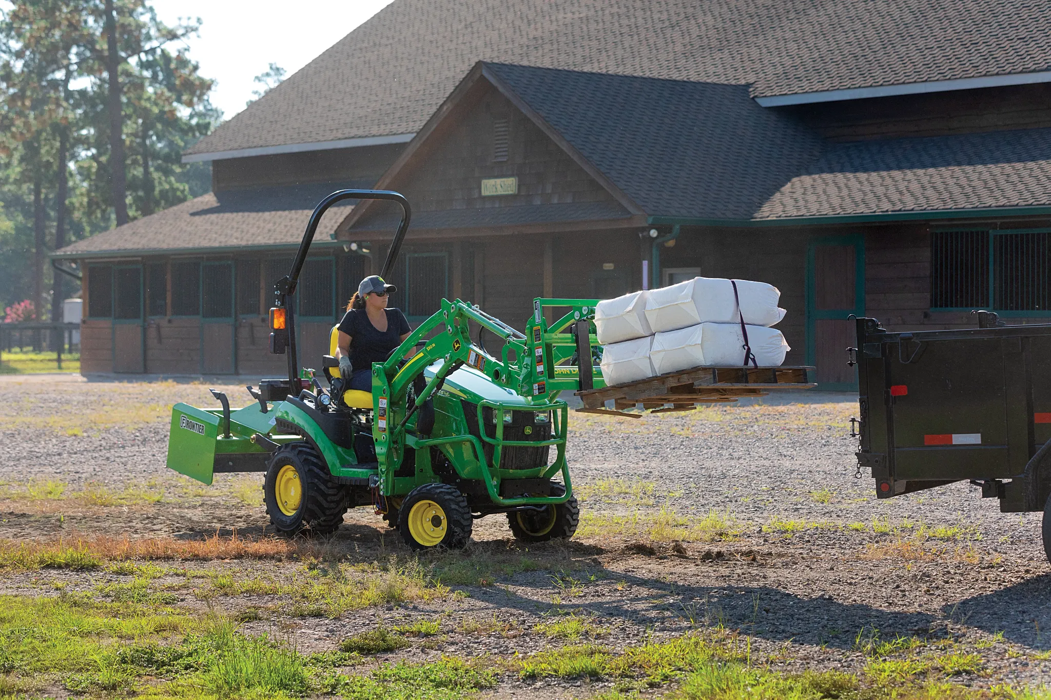 A person operates a small green tractor carrying stacked white bags on the front loader while driving across a gravel area in front of a large horse stable, with a black trailer nearby on a sunny day