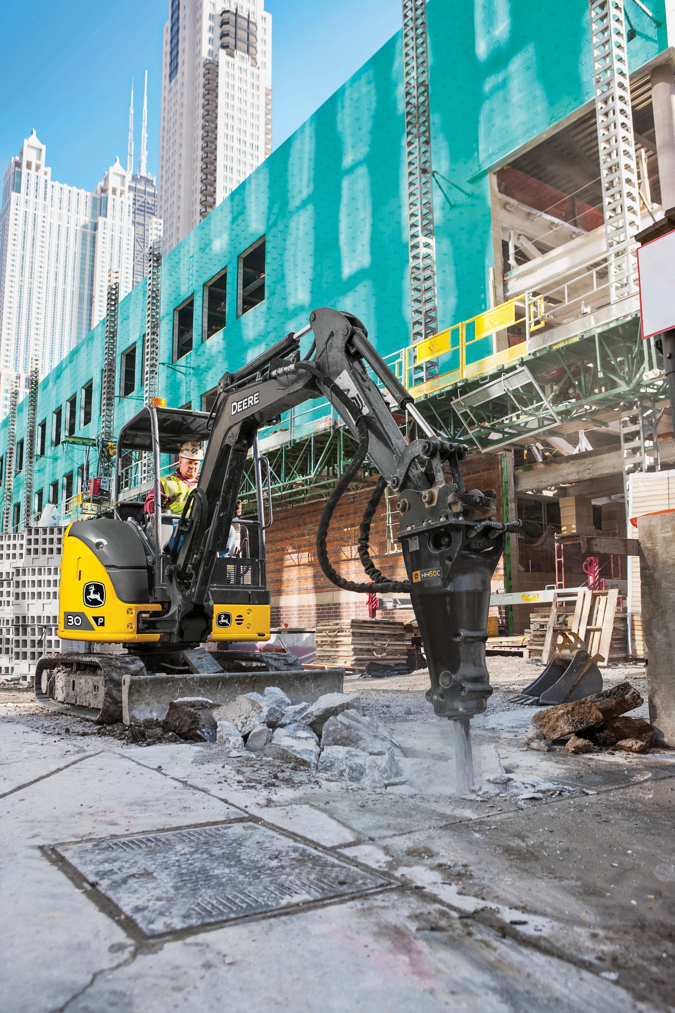 Compact excavator using a hydraulic breaker to jackhammer concrete on a city construction site, with scaffolding and high-rise buildings in the background