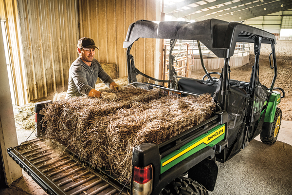 Rear right view of an XUV825M S4 Gator with operator loading the cargo box with hay bales in a barn