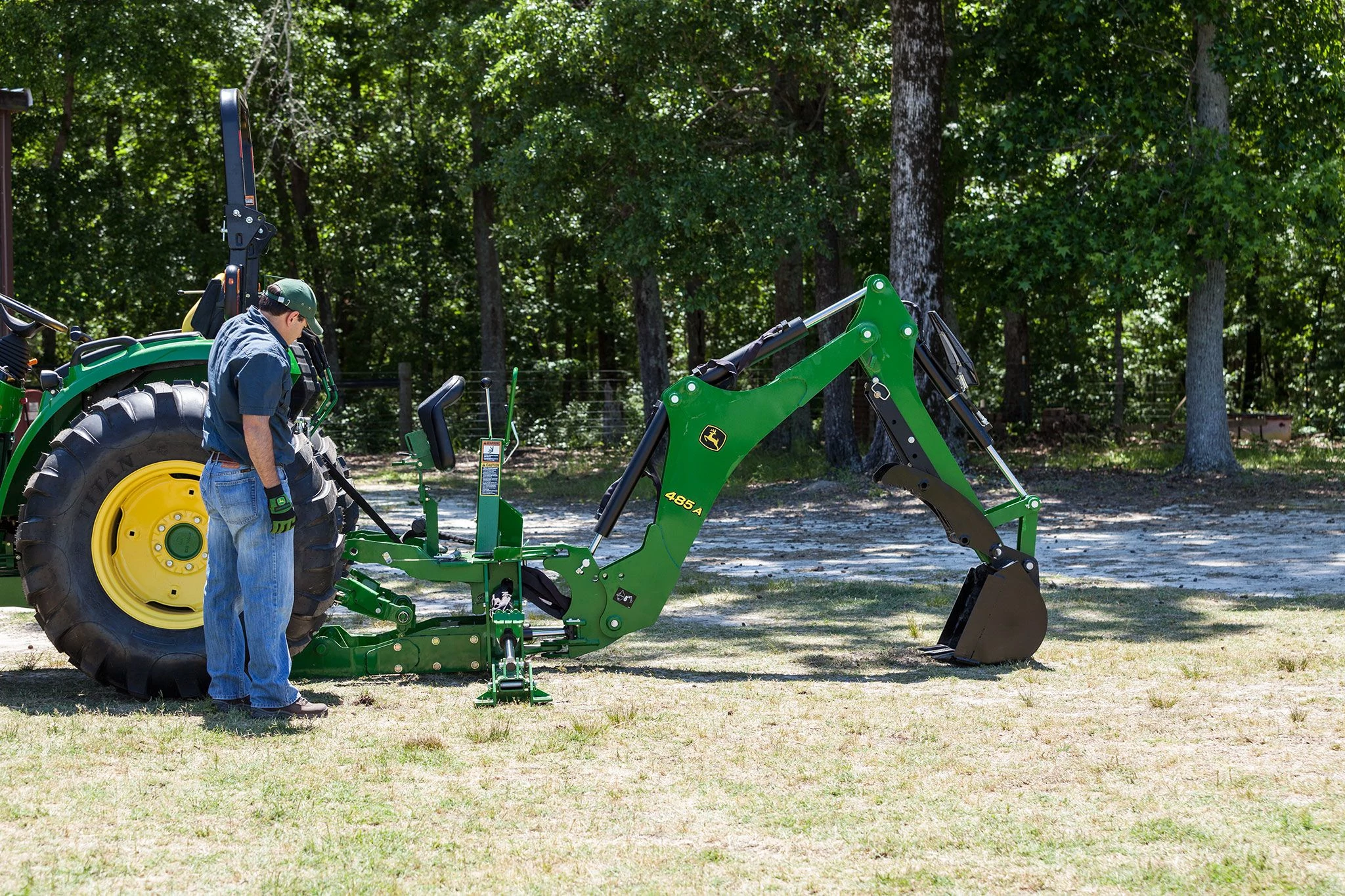 Side view of an operator standing beside a tractor's rear wheel with a 485A backhoe hookup on the back, extended and resting on grass