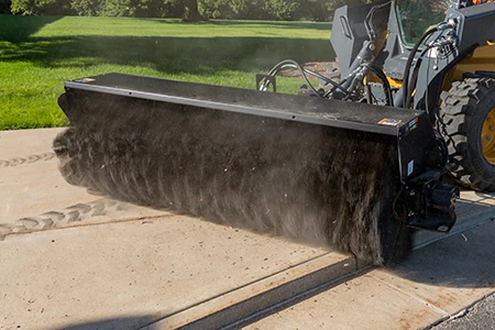 Front left view of a BA96C angle broom brushing a sidewalk with part of the attached skid steer in the background
