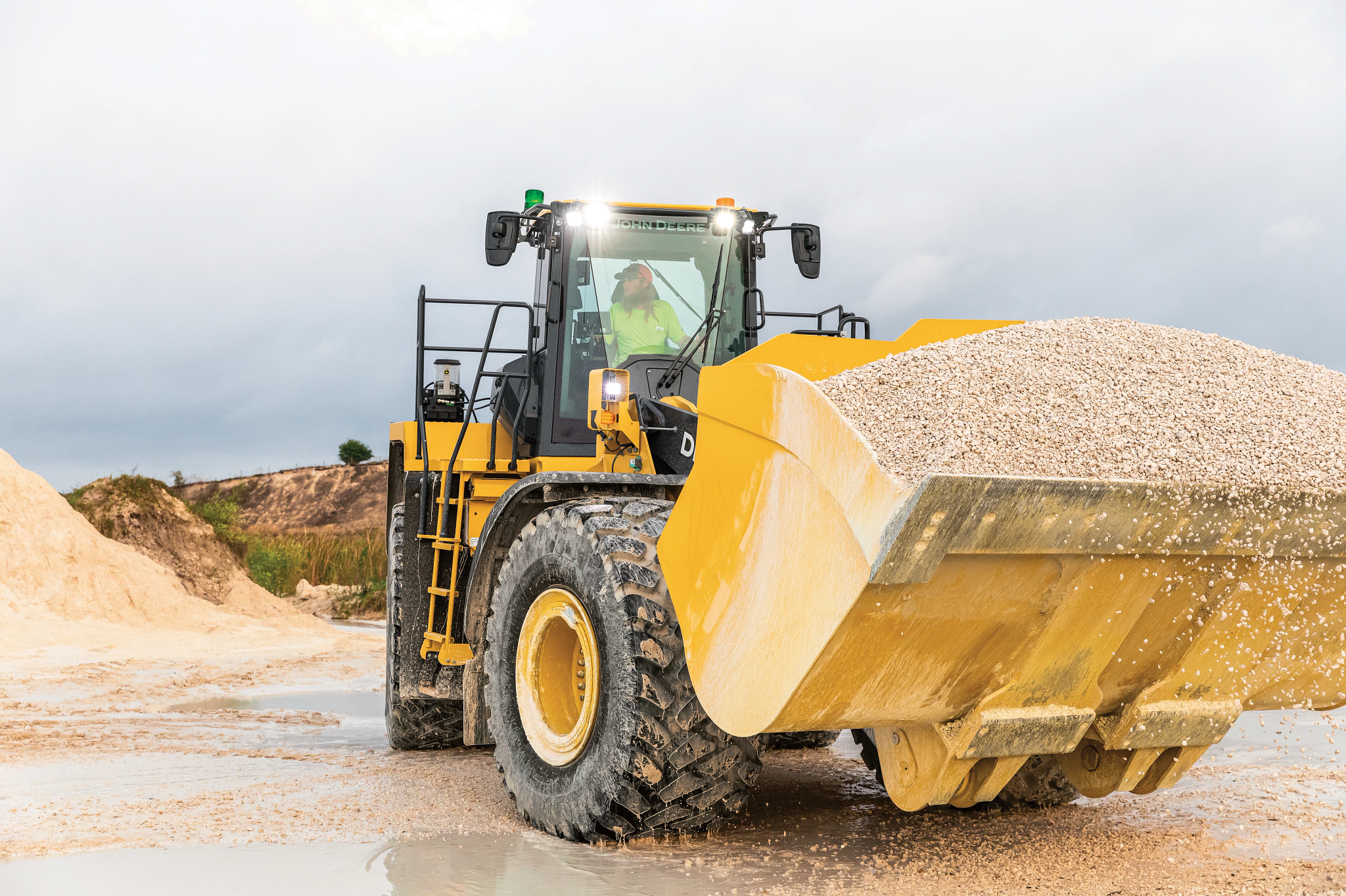 A large yellow front-end loader carries a full bucket of light-colored gravel at a muddy construction site