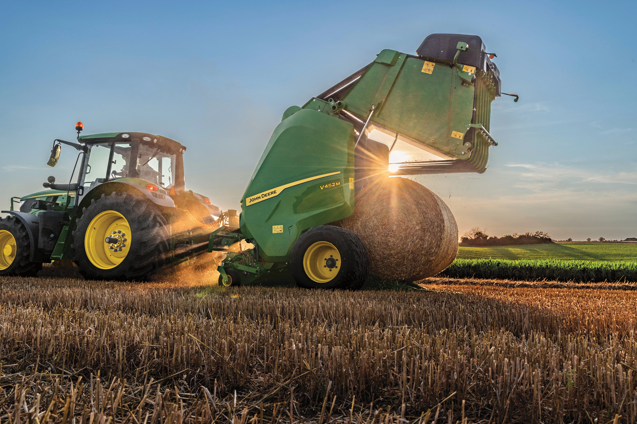 John Deere tractor pulling a green round baler at dusk as a completed round hay bale is discharged onto a harvested field, with dust illuminated by work lights.