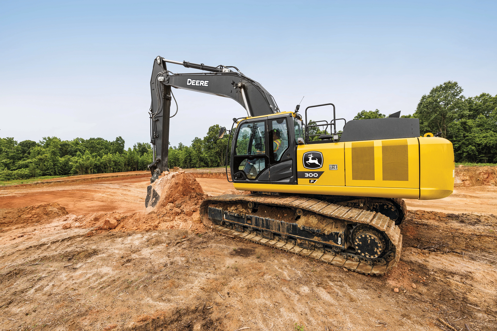 John Deere 350G excavator operating on a dirt construction site, featuring a yellow and gray body, enclosed cab, and metal tracks, with the boom and bucket digging into a mound of soil; surrounded by open earth and a line of green trees under a clear sky