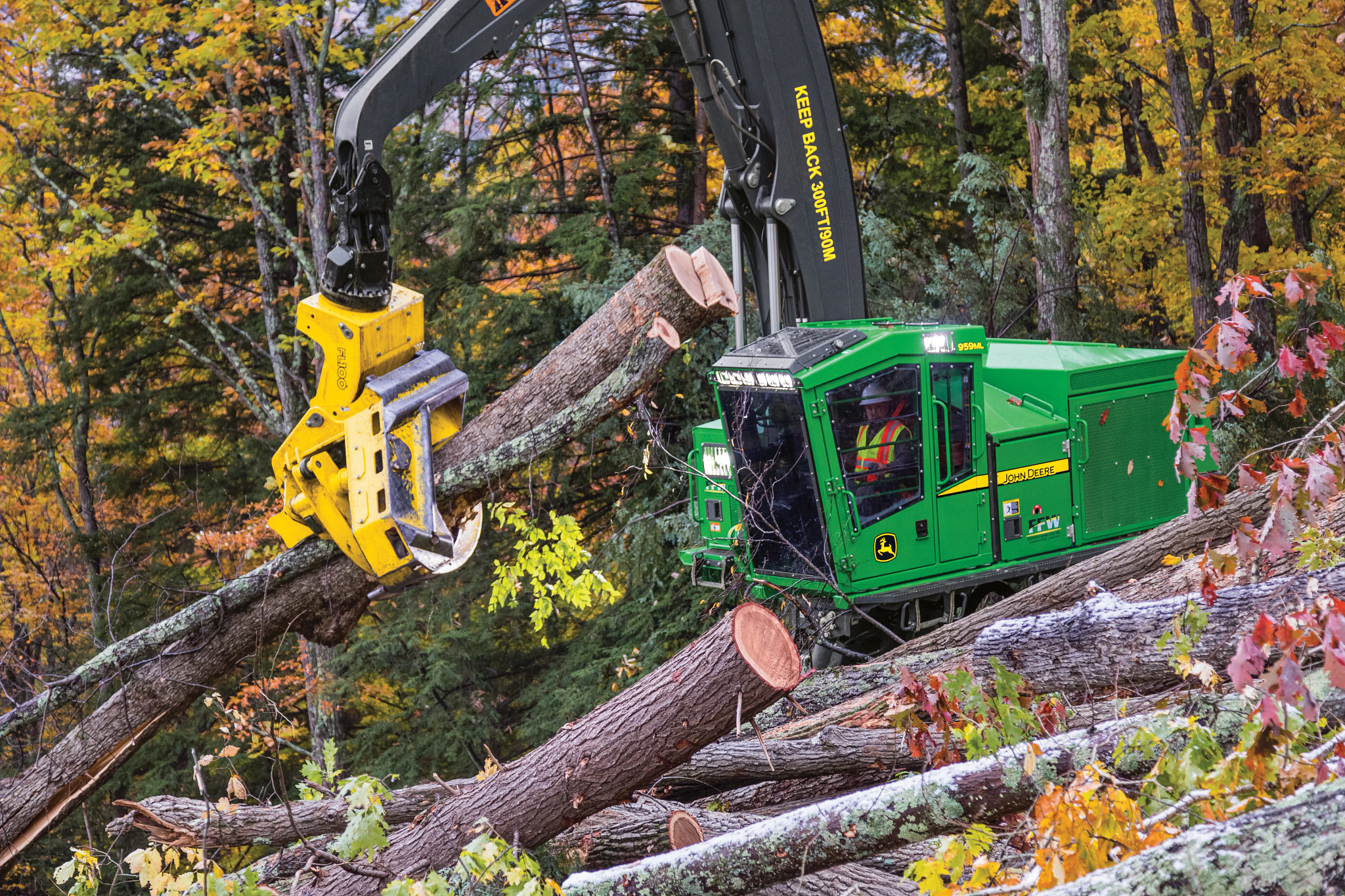 John Deere forestry machine uses a harvesting head to cut and process large logs on a wooded slope, surrounded by fallen timber and trees with autumn foliage in a dense forest setting.