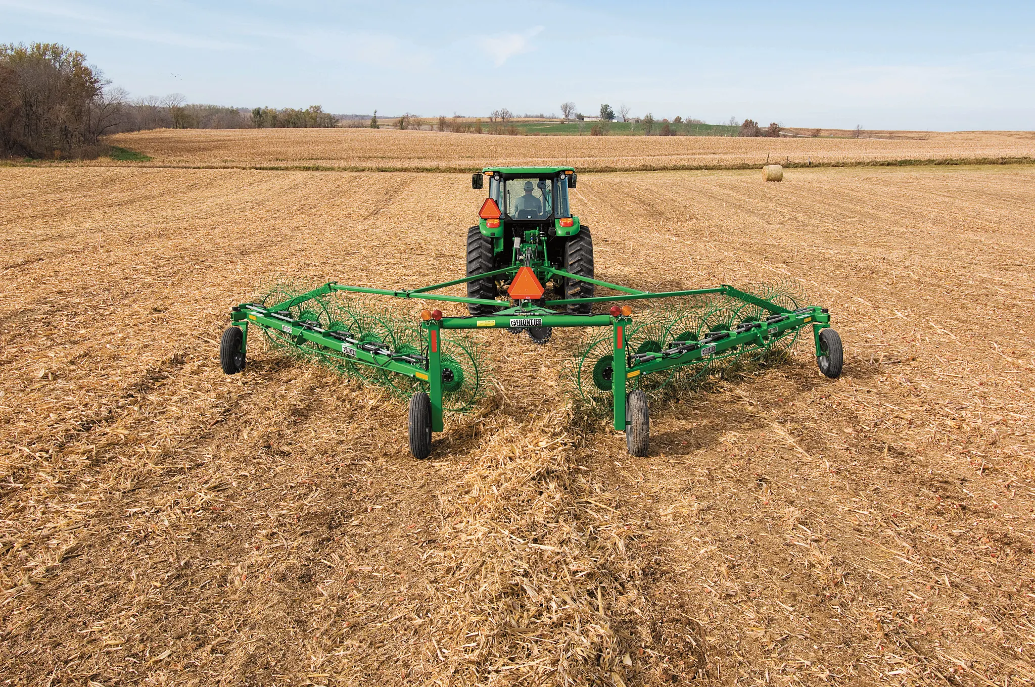 Rear view of a tractor pulling a green WR1212 High-Capacity Wheel Rake across a tan, harvested field