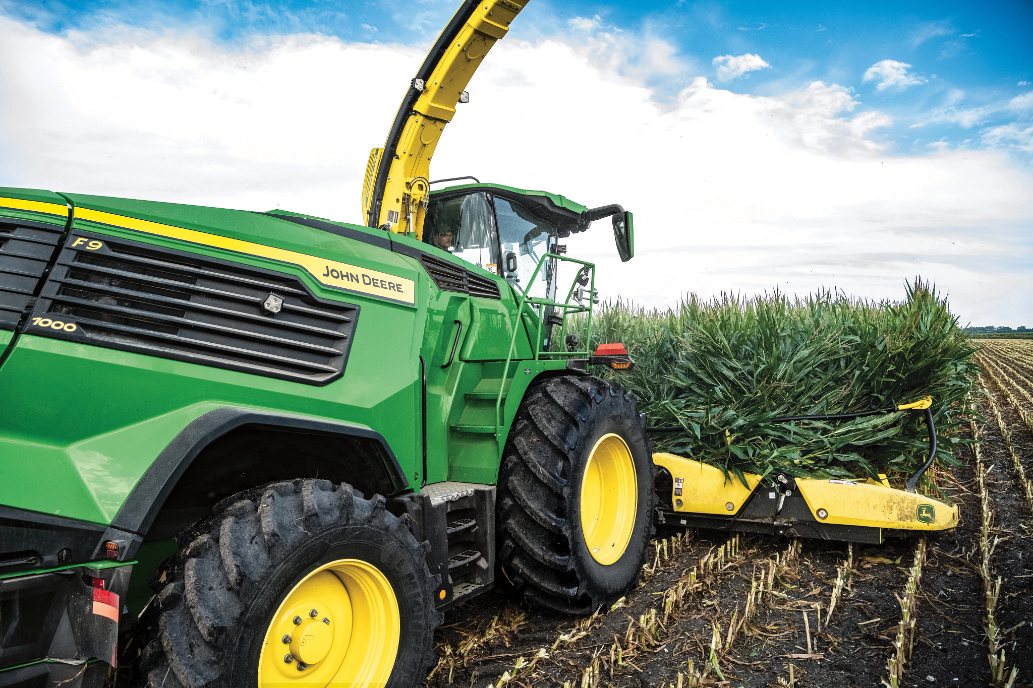 John Deere F9 7000 self‑propelled forage harvester harvesting corn silage, shown from front‑left with yellow corn head, large front tires, and green body working through a field under blue sky.