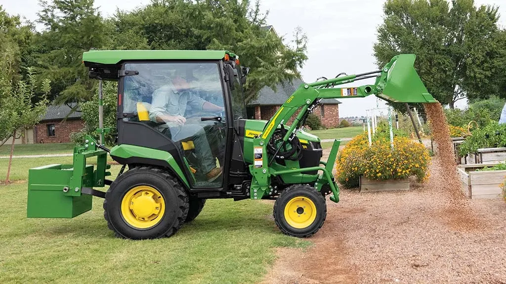 A green compact tractor with a rear ballast box and front loader dumps gravel around some planter boxes.