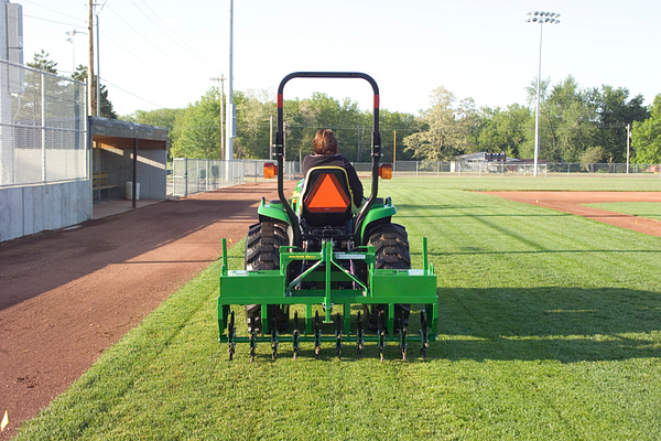 Rear view of a John Deere compact tractor with rear-attached core aerator traveling in a straight line on a baseball field
